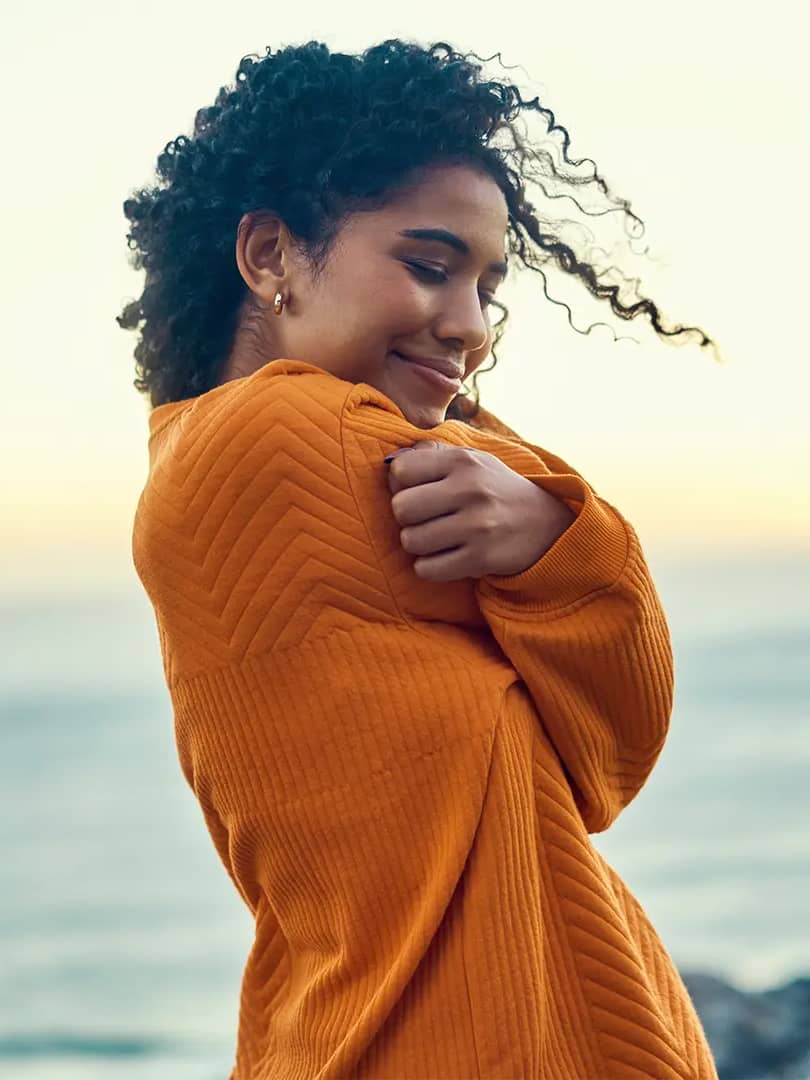 Titelbild zum Artikel "Selbstliebe & Affirmation": Selbstbewusste Frau umarmt sich selbst am Meer