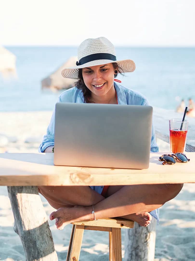 Titelbild für Artikel "Workation": Frau sitzt am Strand vor dem Meer und arbeitet am Laptop