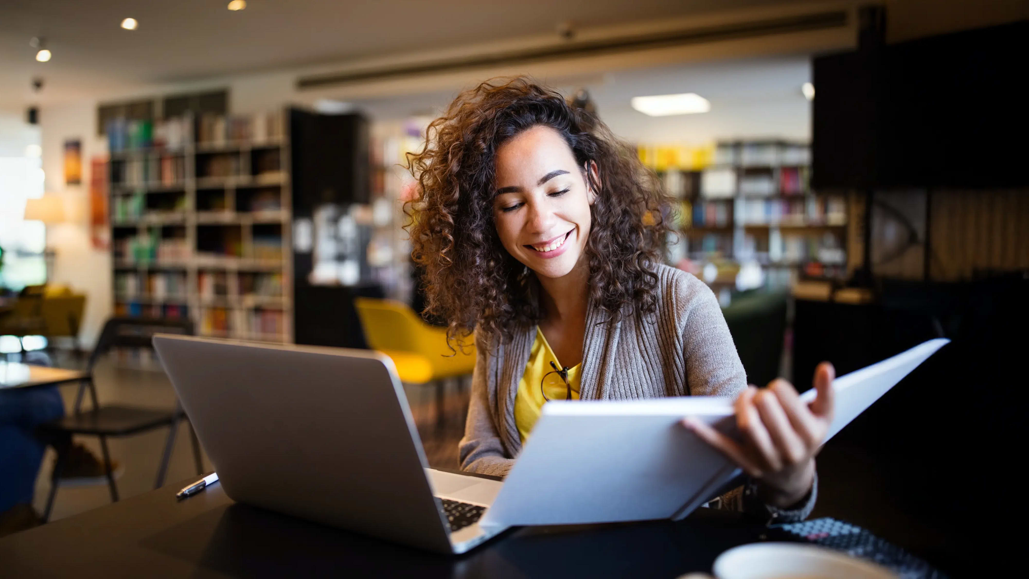 Eine Frau sitzt lächelnd an einem Tisch mit einem Laptop und Dokumenten in einer gemütlichen Bibliothek oder einem Café. Die Umgebung mit Bücherregalen und bequemen Sitzmöglichkeiten schafft eine produktive und einladende Atmosphäre.