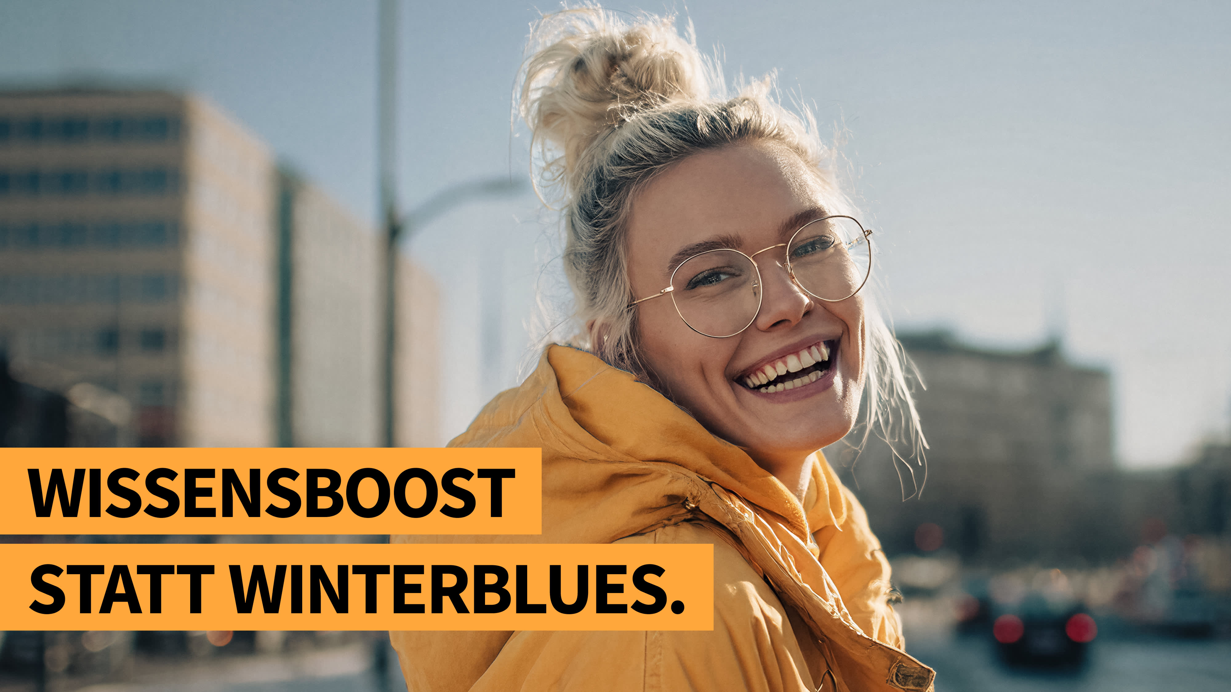 Lachende junge Frau mit Brille und gelbem Parka in städtischer Umgebung bei Sonnenlicht