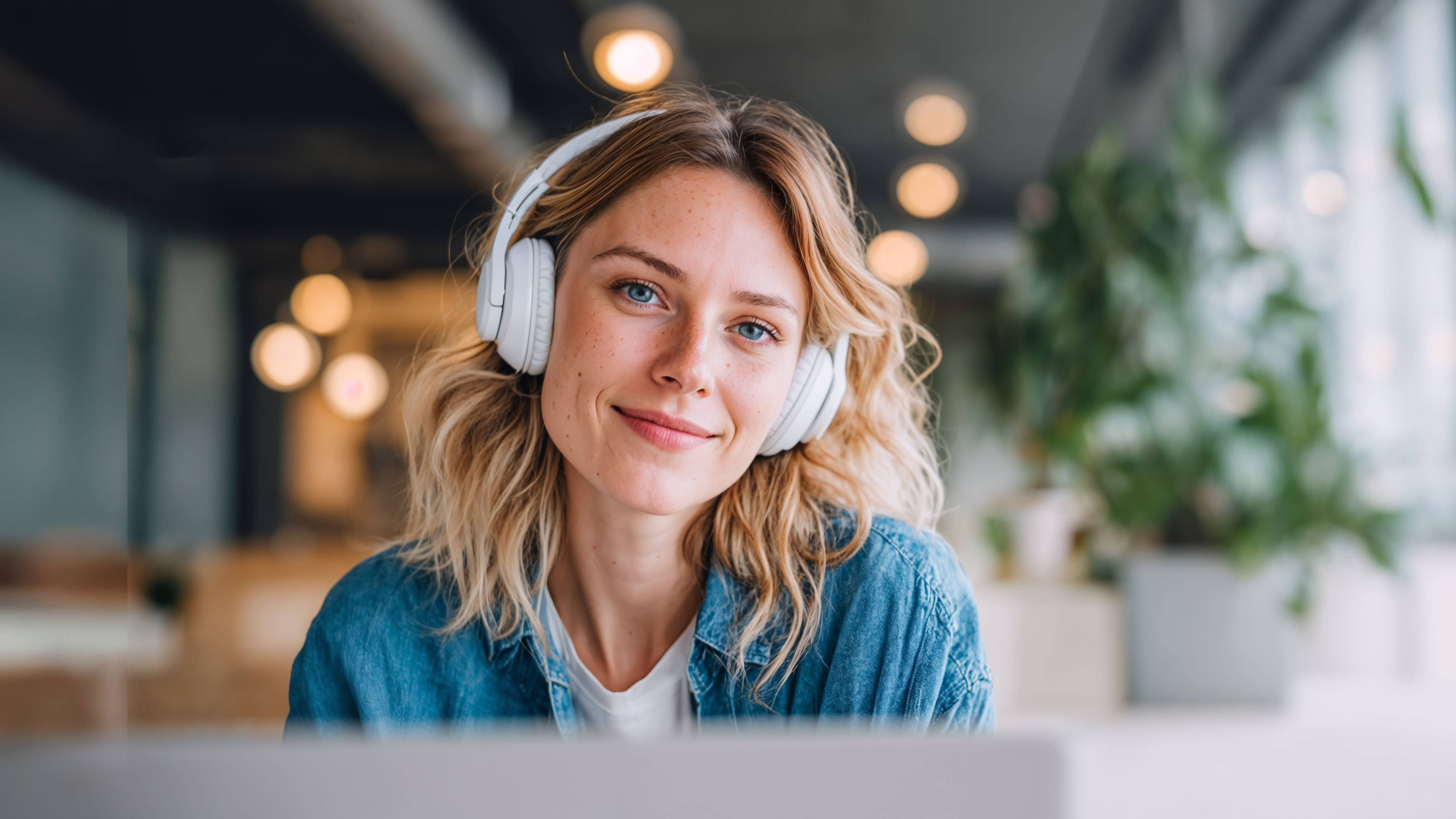 Junge blonde Frau mit weißen Over-Ear-Kopfhörern und Jeansjacke sitzt lächelnd vor einem Laptop in einem modernen, hellen Büro.