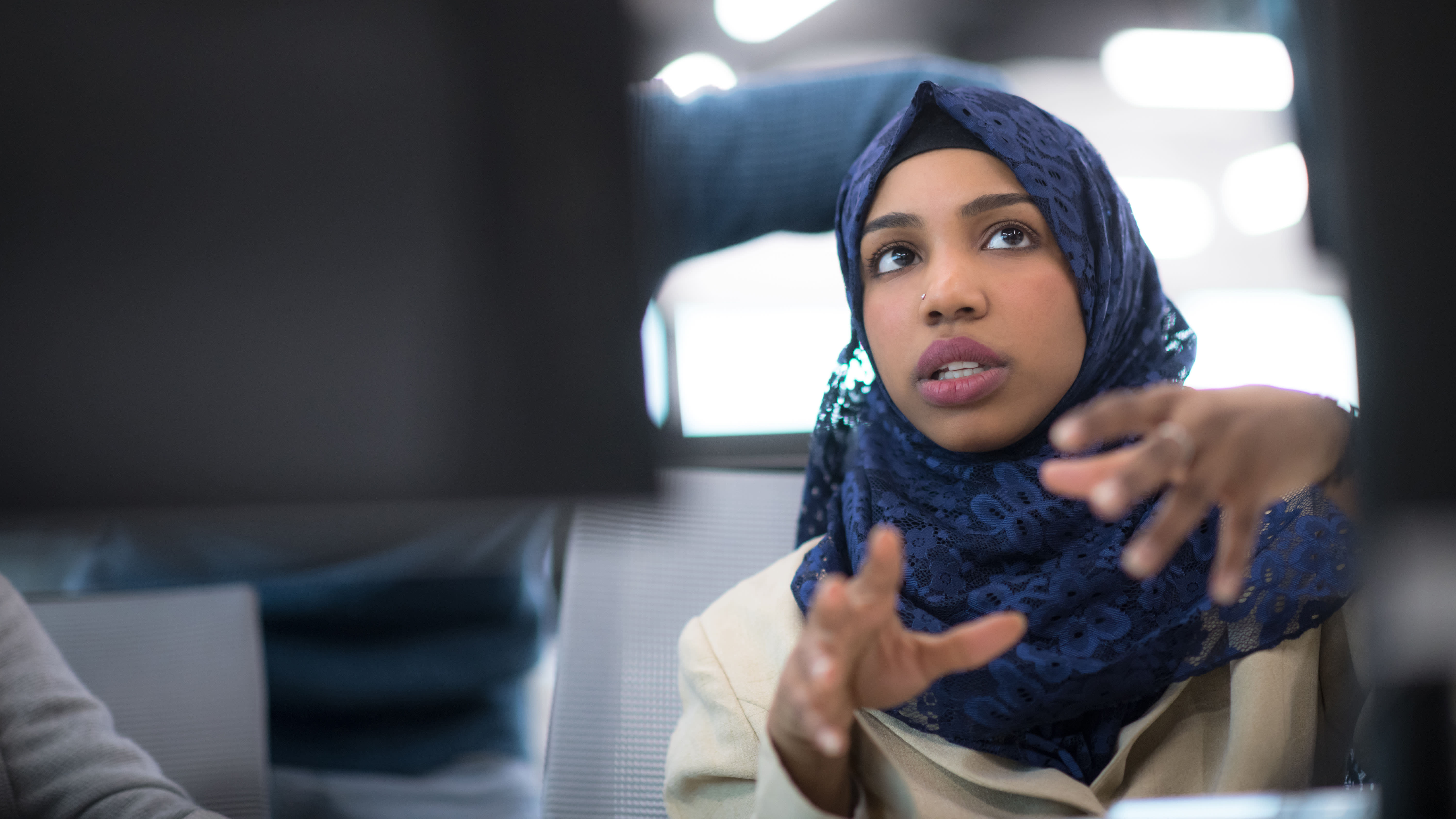 Person in blue lace headscarf gestures with both hands while speaking; another person in striped shirt is blurred in the background. Indoors setting.
