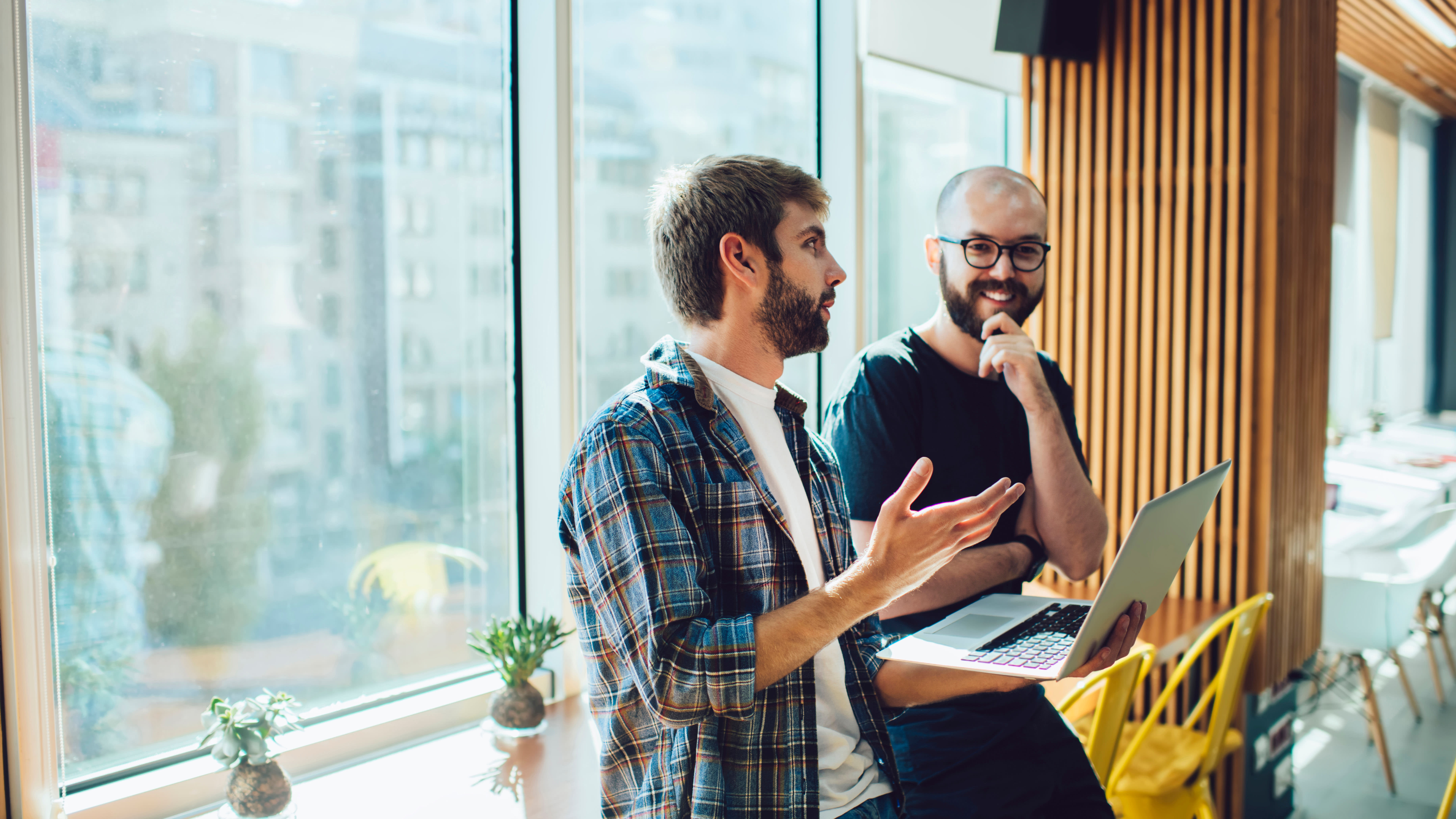 Two people in a modern office discuss something; one holds a laptop, the other listens with hand on chin. Yellow chairs and plants in background.