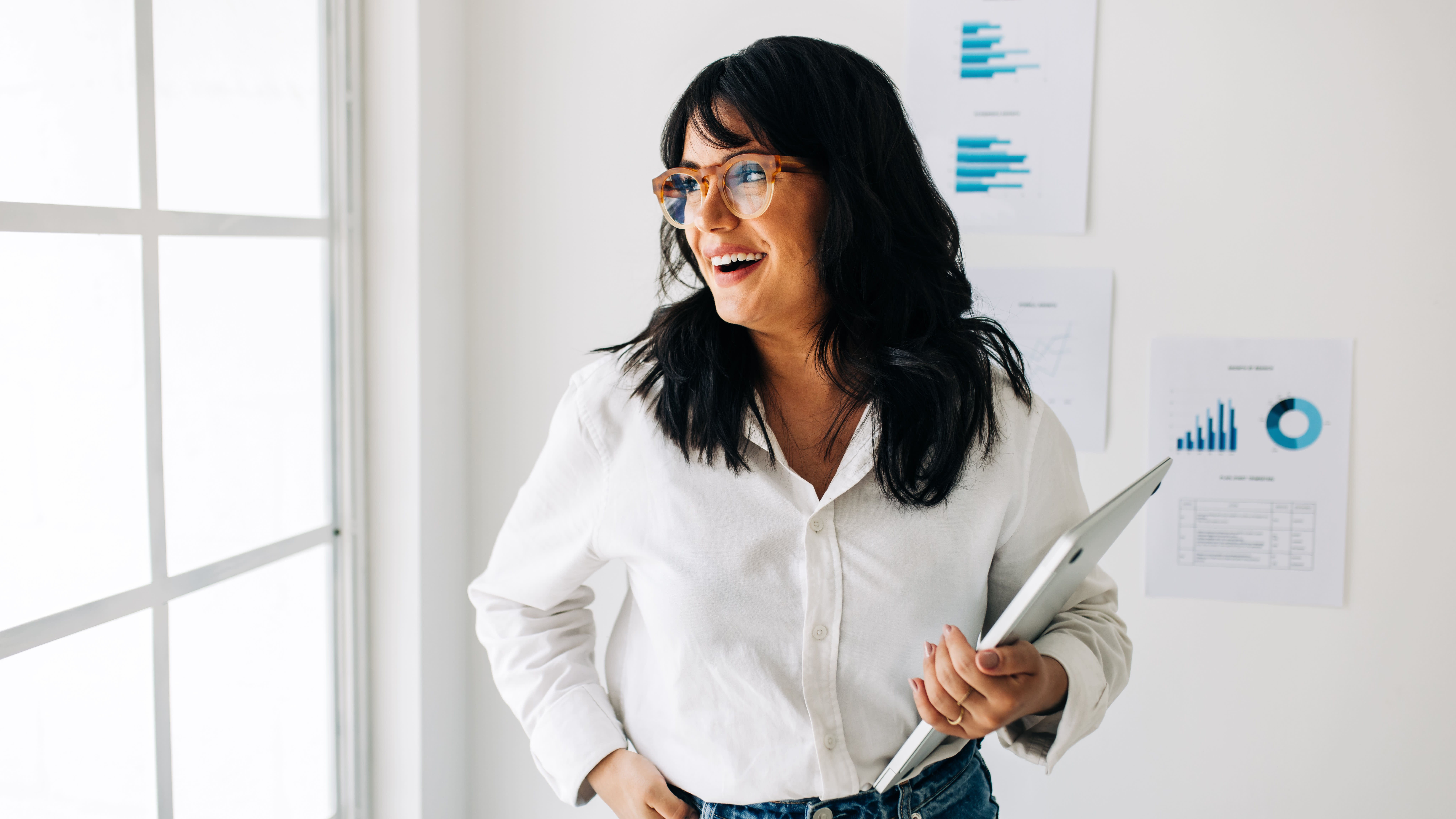 Woman in glasses and white shirt smiles while holding a laptop, standing by charts on a bright office wall.