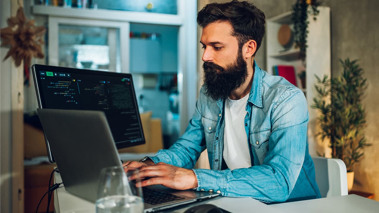 A person in a denim shirt works on advanced data analysis in a home office, using a laptop and a secondary screen displaying code.