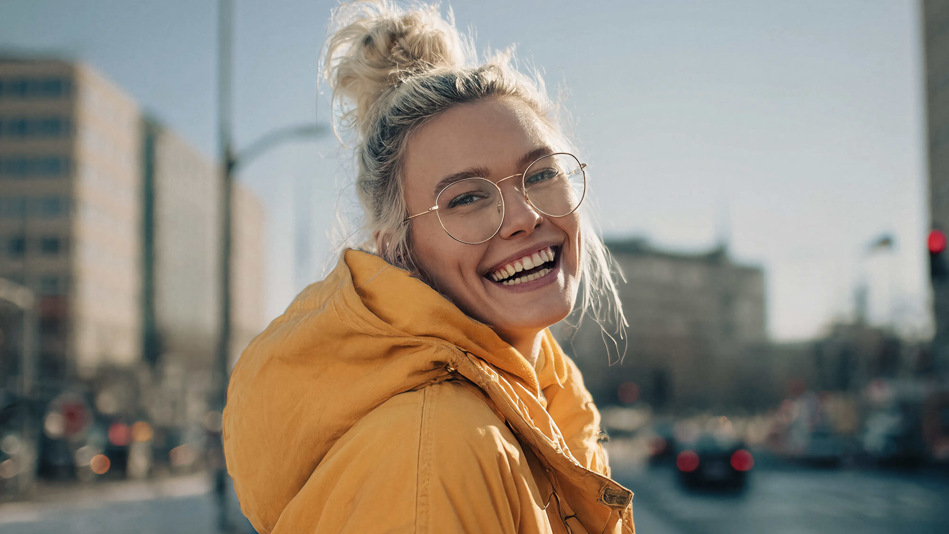 Lachende junge Frau mit Brille und gelbem Parka in städtischer Umgebung bei Sonnenlicht