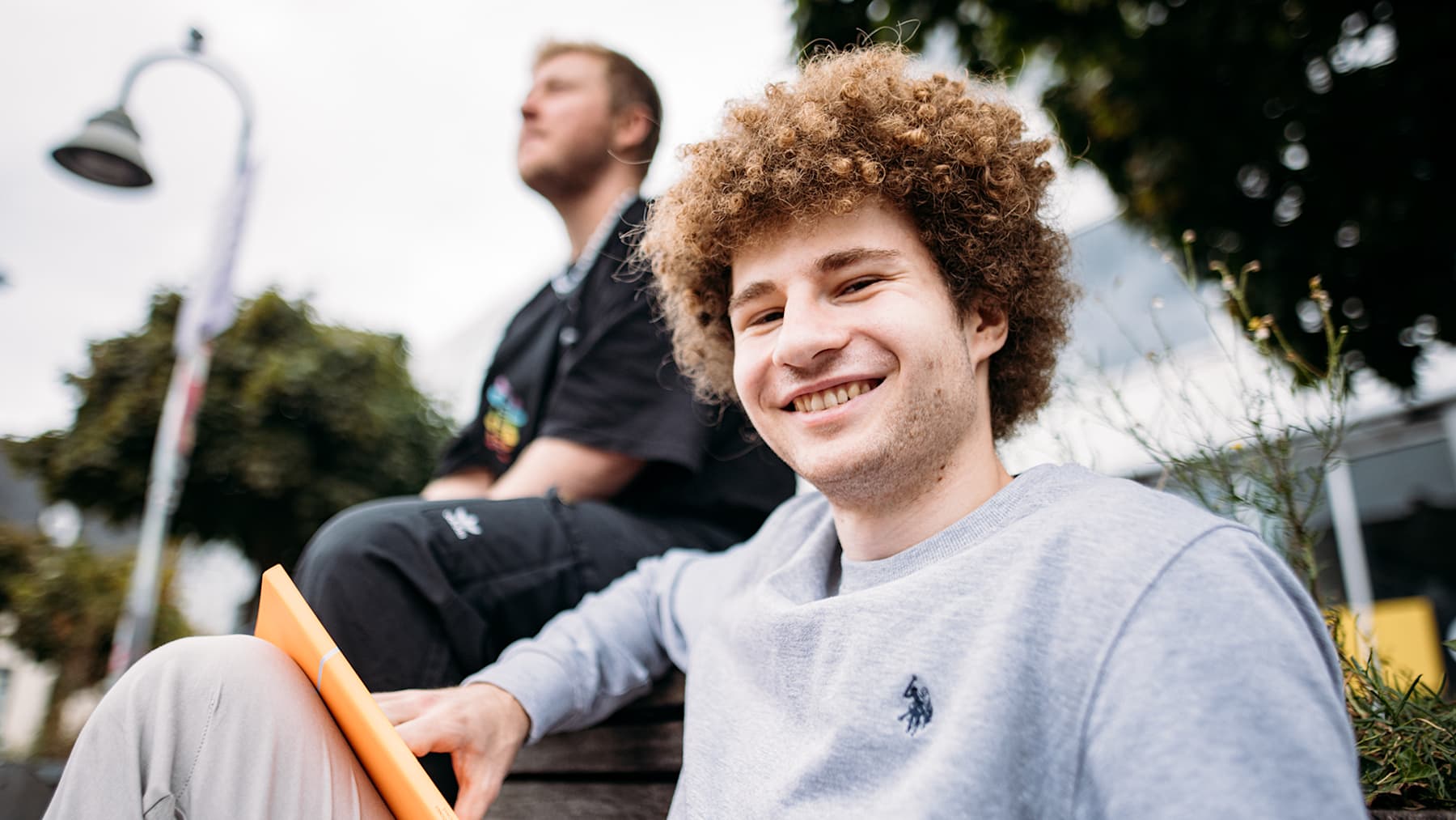 A young guy with curly hair is smiling at the camera, holding an orange IU notebook.