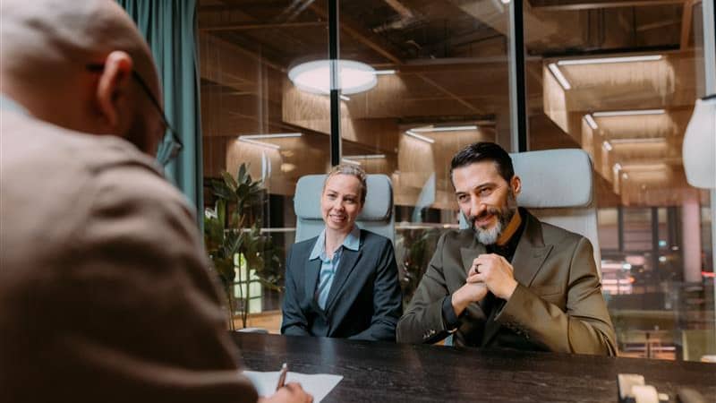 Two employers sit and smile to a new employee who sings a job contract in an office.