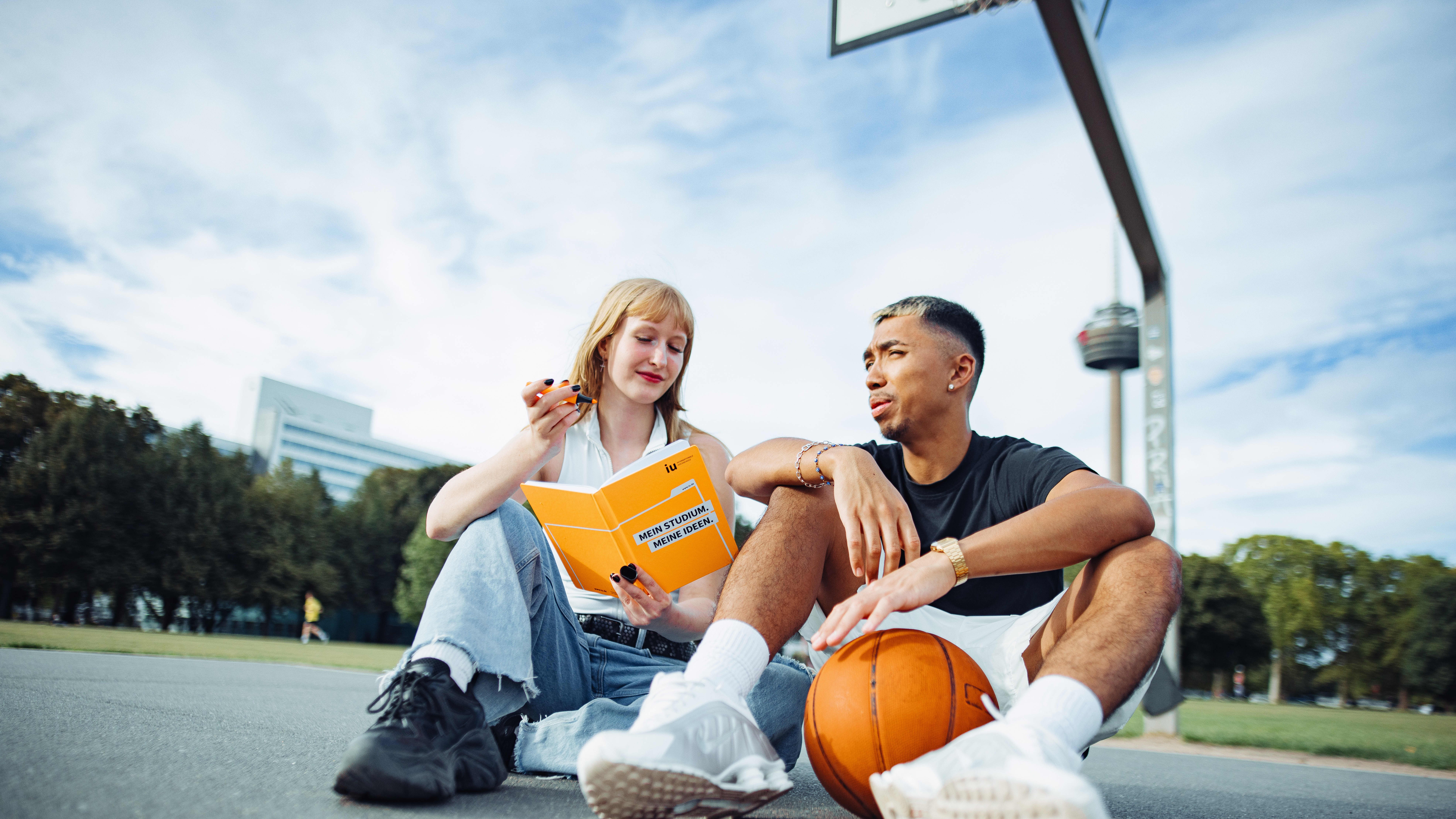 A couple sits on a basketball court. She is holding an IU notebook, and he is holding a basketball.