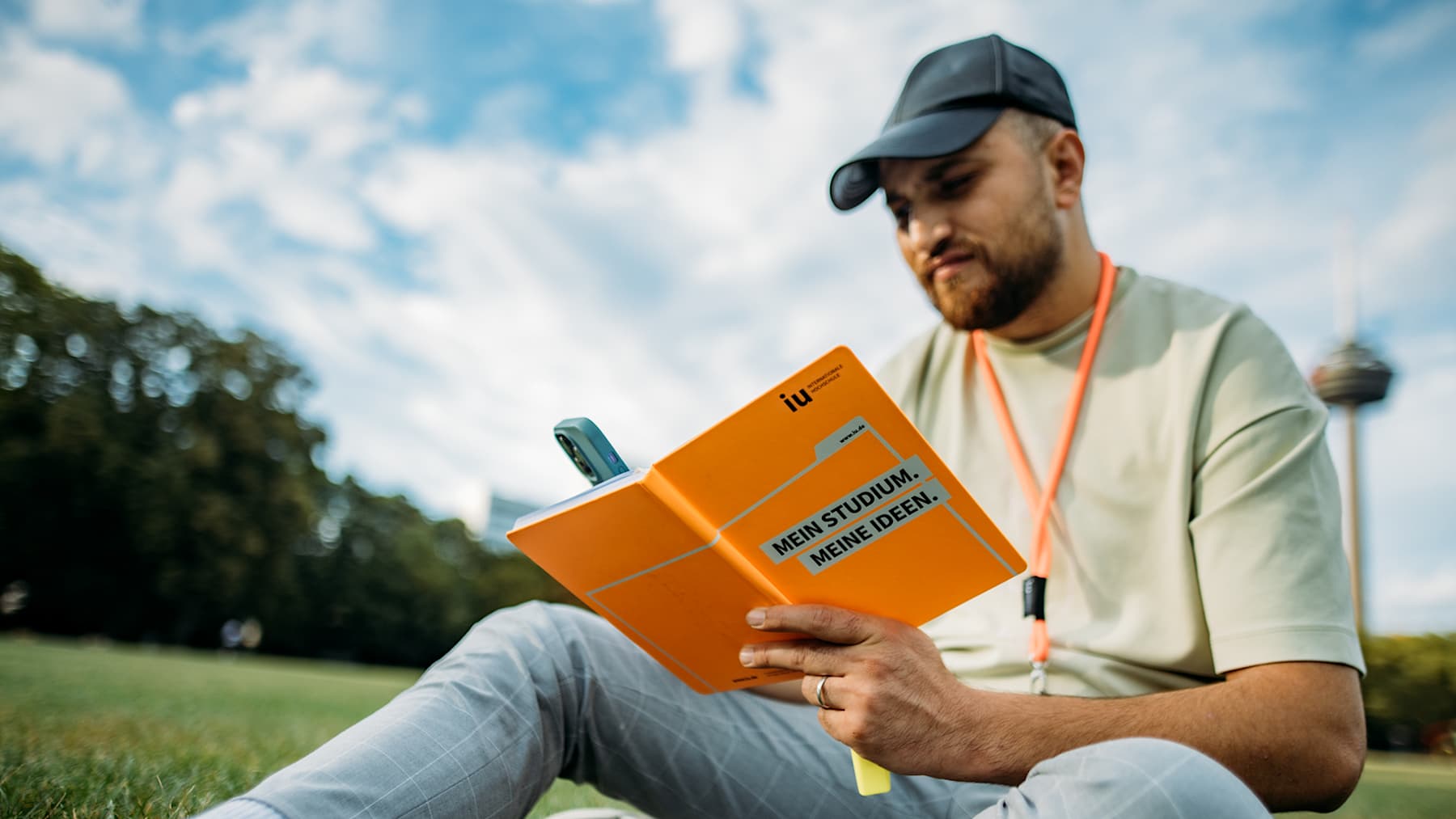 A person wearing a baseball cap and a light green t-shirt sits on grass and reads an orange IU notebook with "MEIN STUDIUM. MEINE IDEEN." written on it.