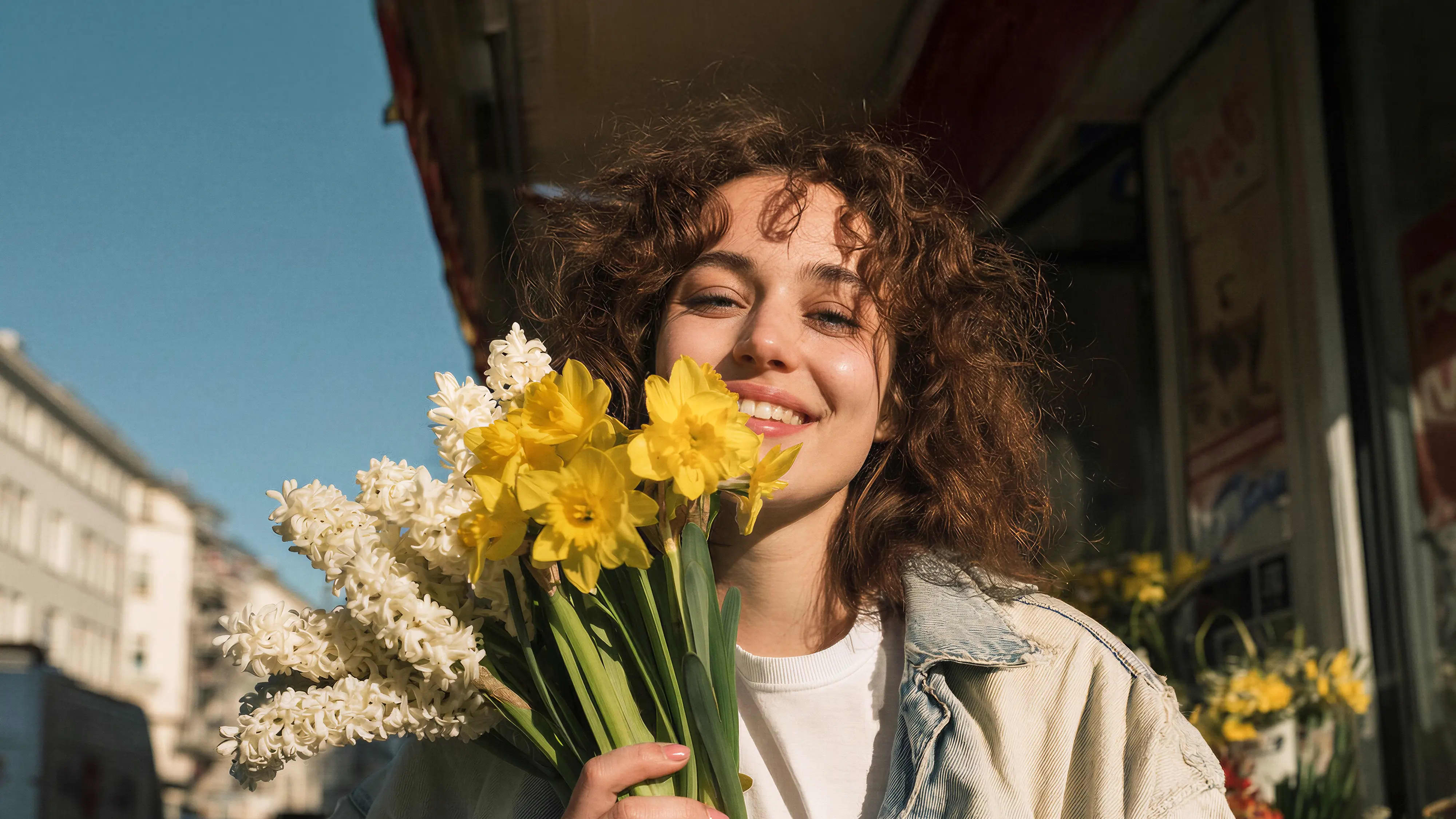 Lächelnde Frau mit lockigem Haar hält einen Strauß gelber und weißer Frühlingsblumen vor städtischem Hintergrund