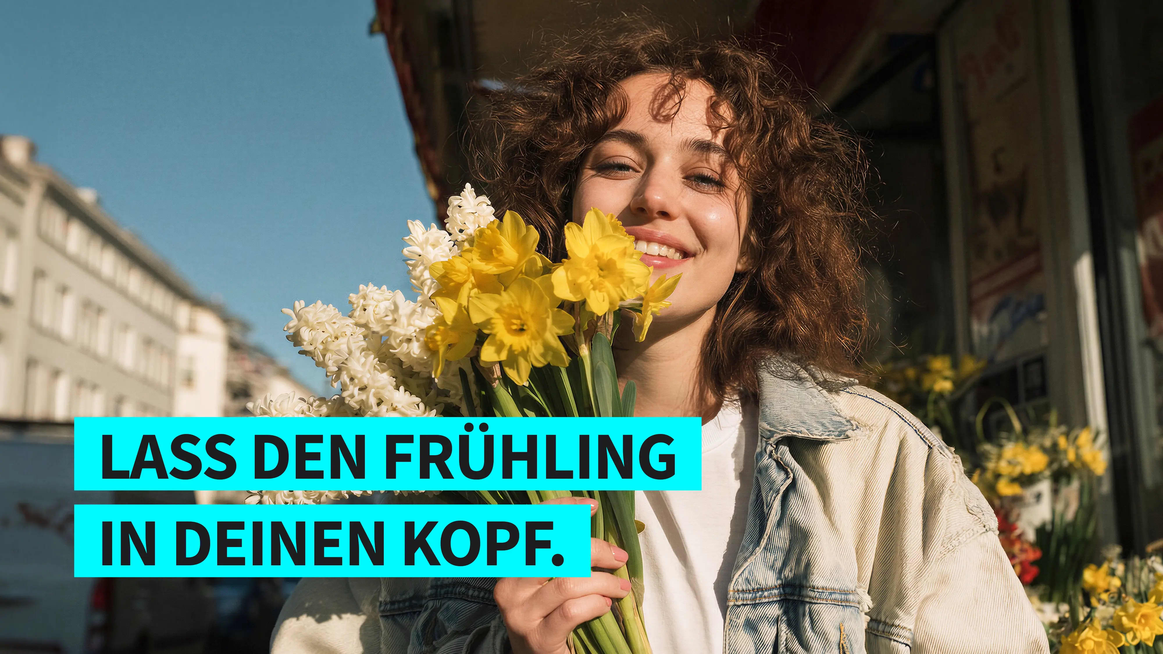 Lächelnde Frau mit lockigem Haar hält einen Strauß gelber und weißer Frühlingsblumen vor städtischem Hintergrund