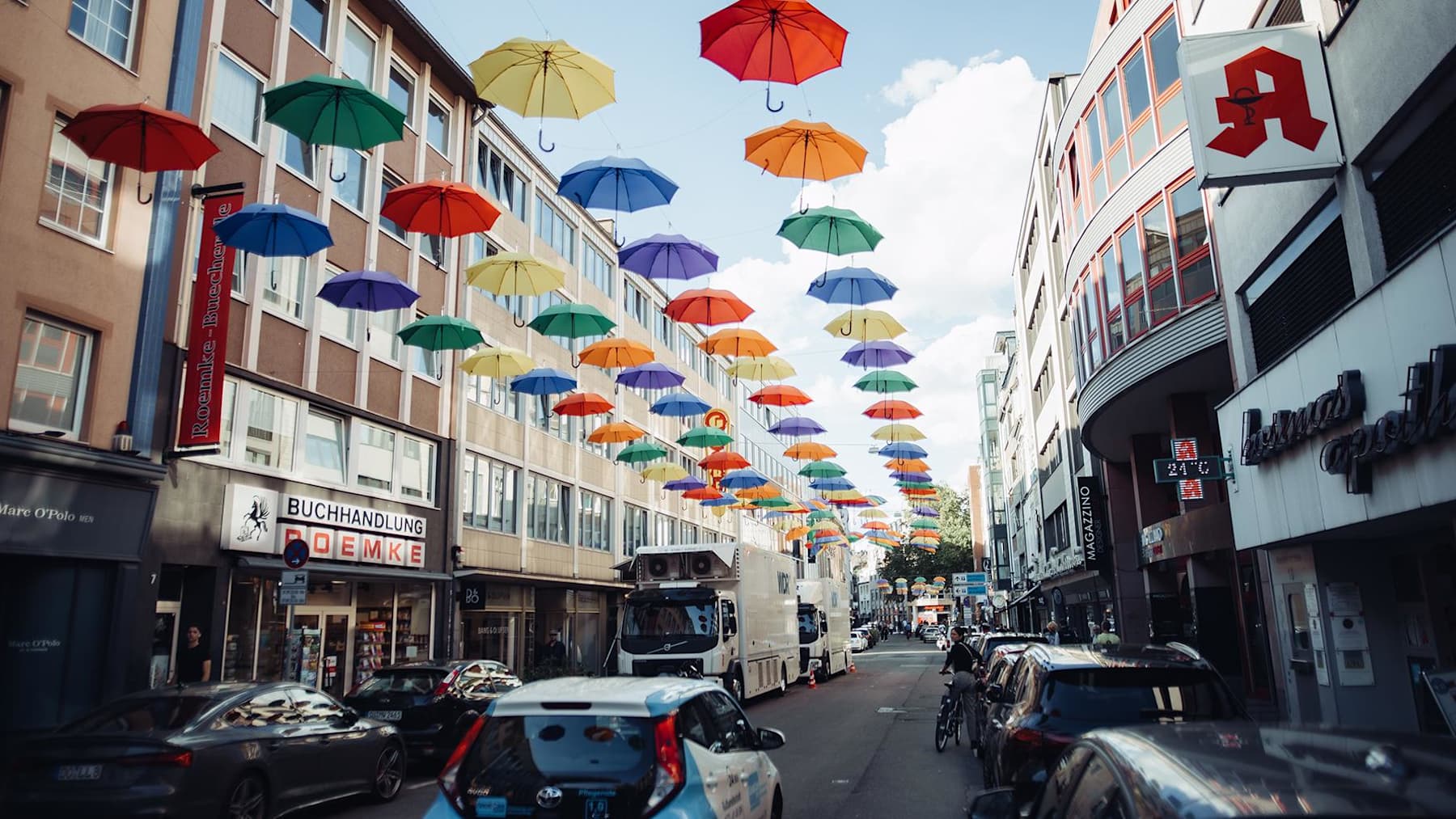 Cologne city street with colorful umbrellas hanging above, cars parked along both sides, and people walking on the sidewalks.