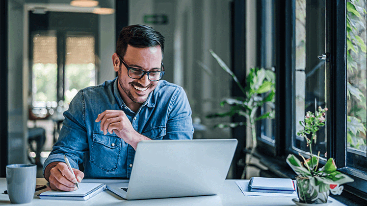 Smiling man at laptop in denim shirt reviewing jobs for various departments as a human resource manager.