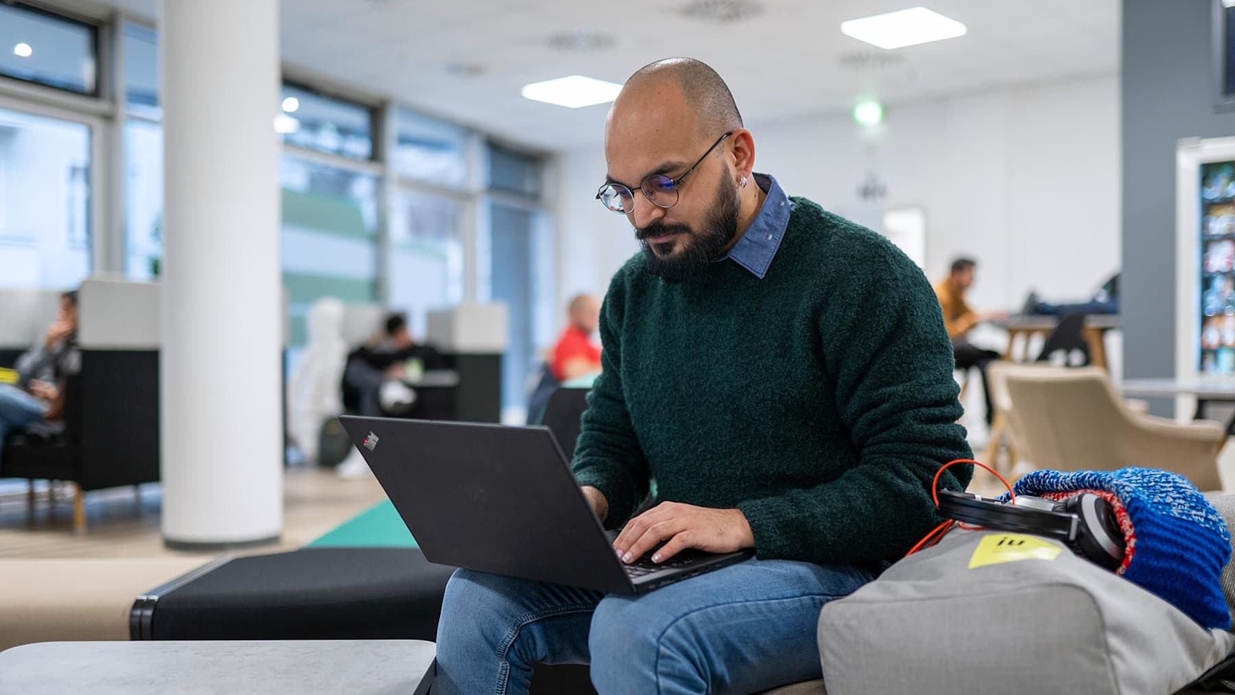 A male student wearing glasses and a beard sits on campus working on a laptop while wearing a green jumper and jeans. An IU-branded bag containing headphones is next to him.