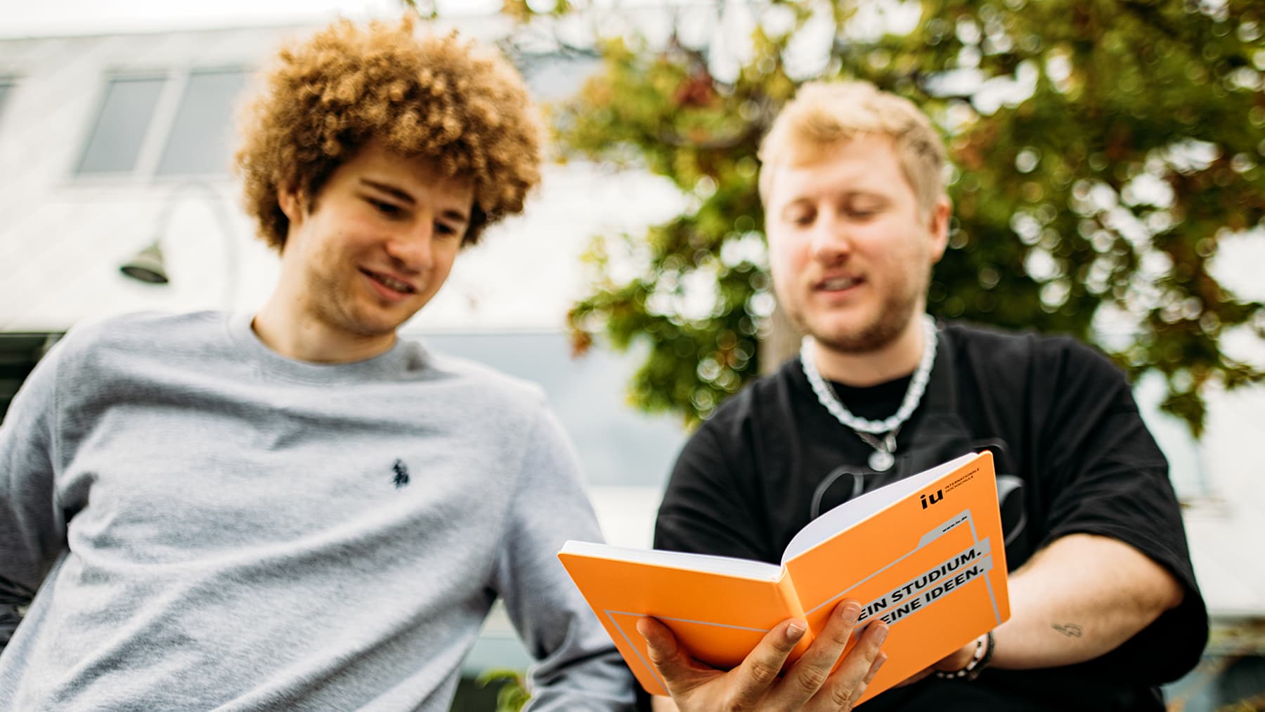Two young students are outside looking at an open, orange, IU-branded notebook.