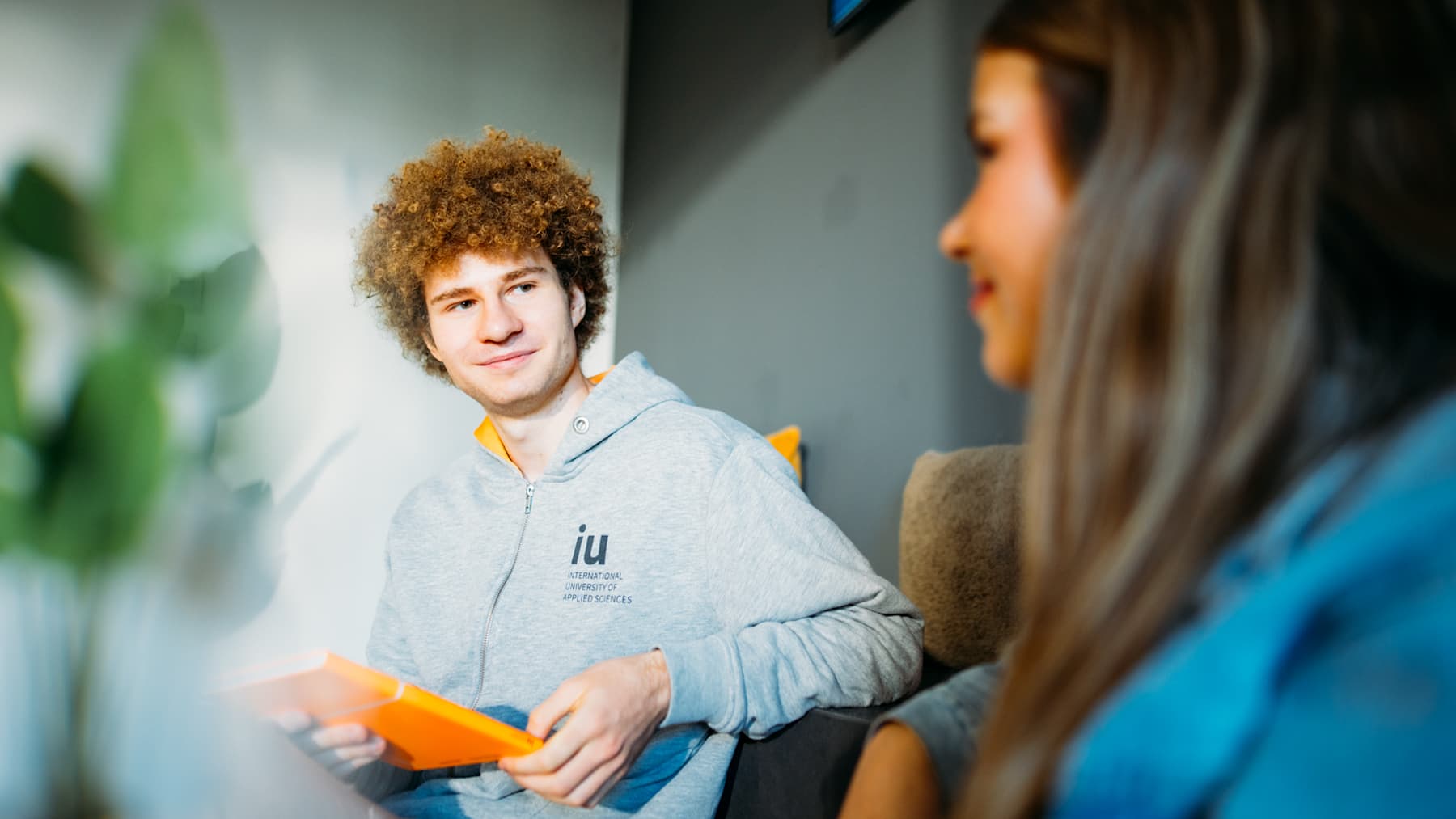Male student at a German university holding a tablet and discussing coursework with a classmate indoors.