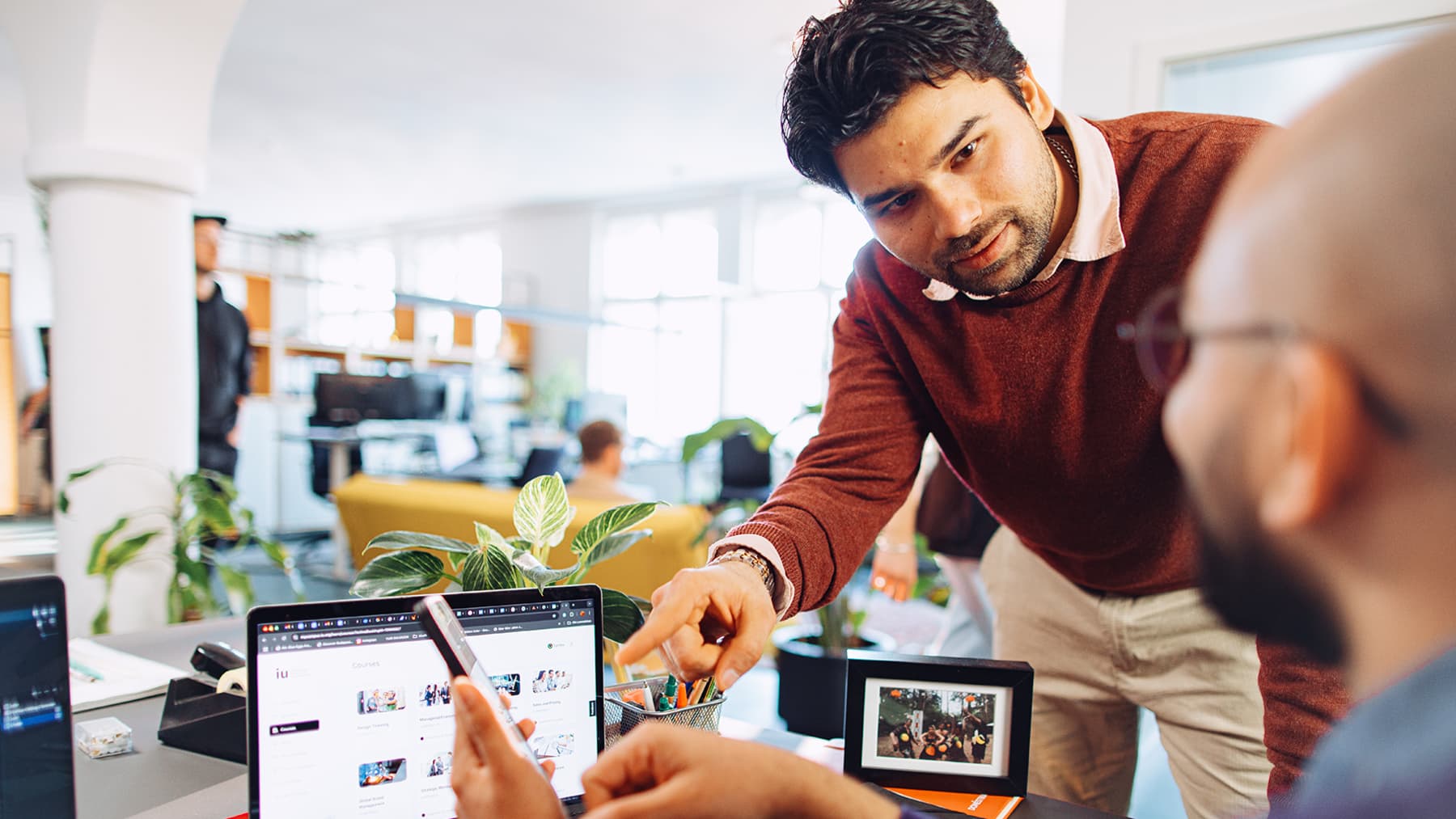 Man with a red sweater is asking for job tips to another man who is using his phone and laptop