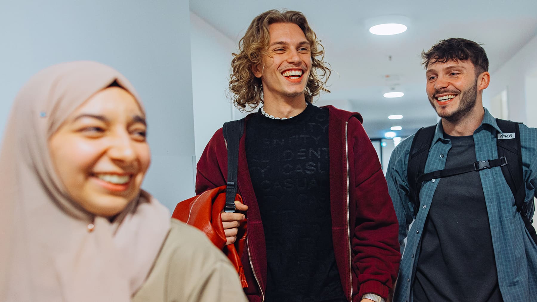 A girl wearing a headscarf and two boys carrying their backpacks are laughing and walking together in a university corridor.