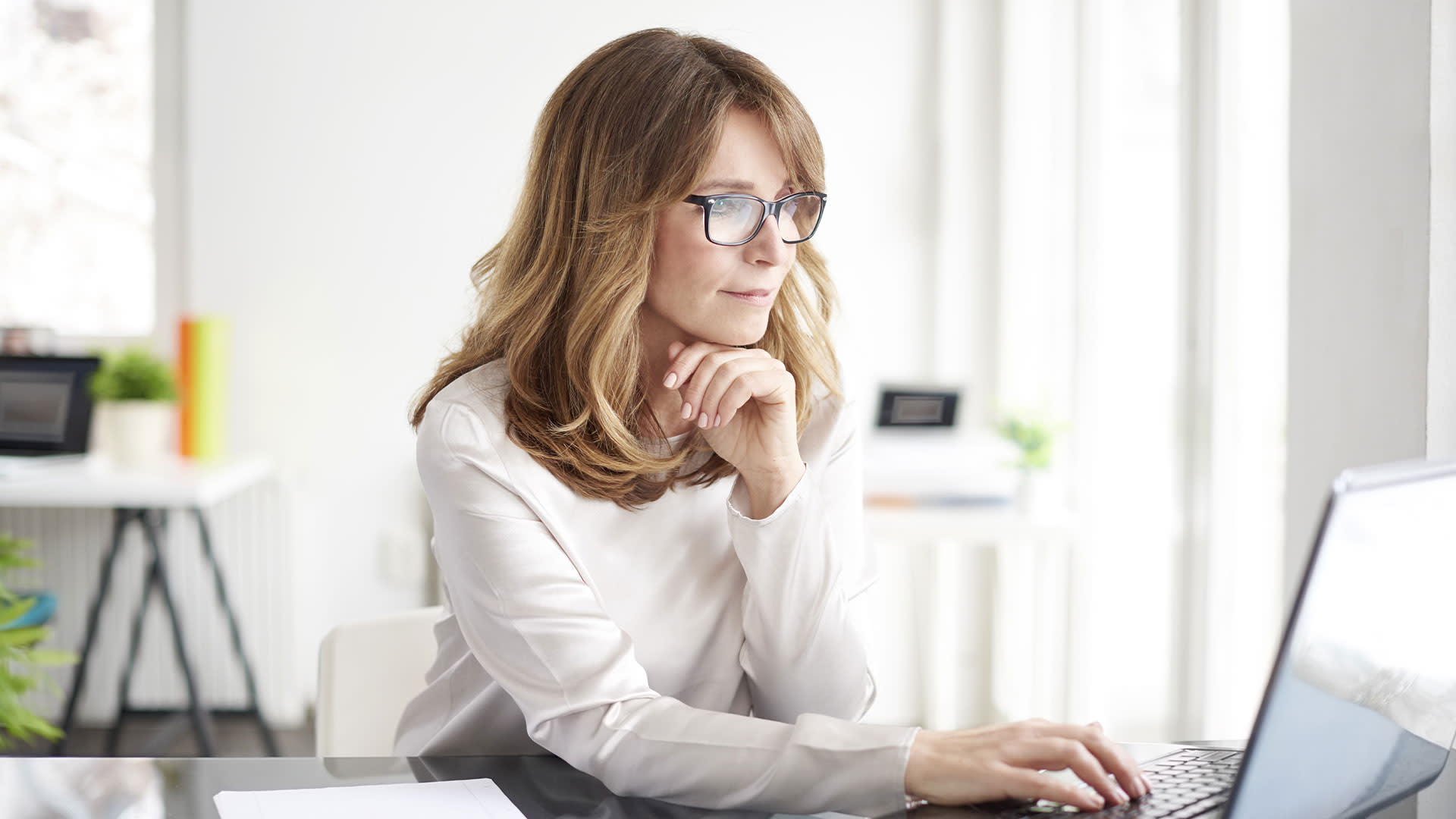 A woman wearing glasses sitting at her desk looking at her laptop.