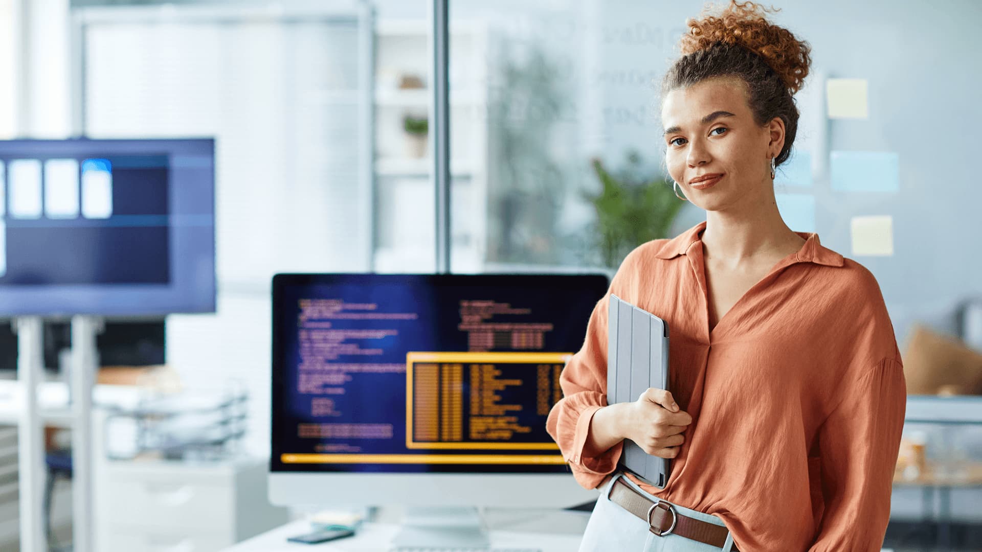 Curly hair woman wearing an orange shirt is holding her ipad which she uses to expertly analyse data