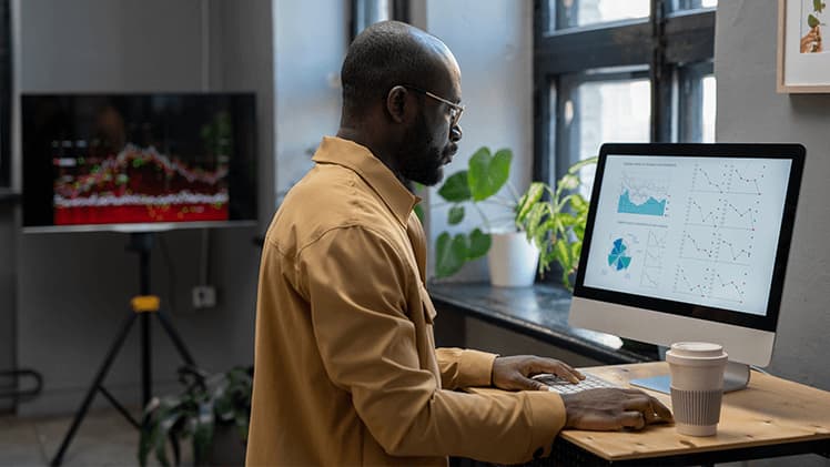 Guy wearing glasses and orange shirt is sitting in front of his PC analysing data sets and patterns