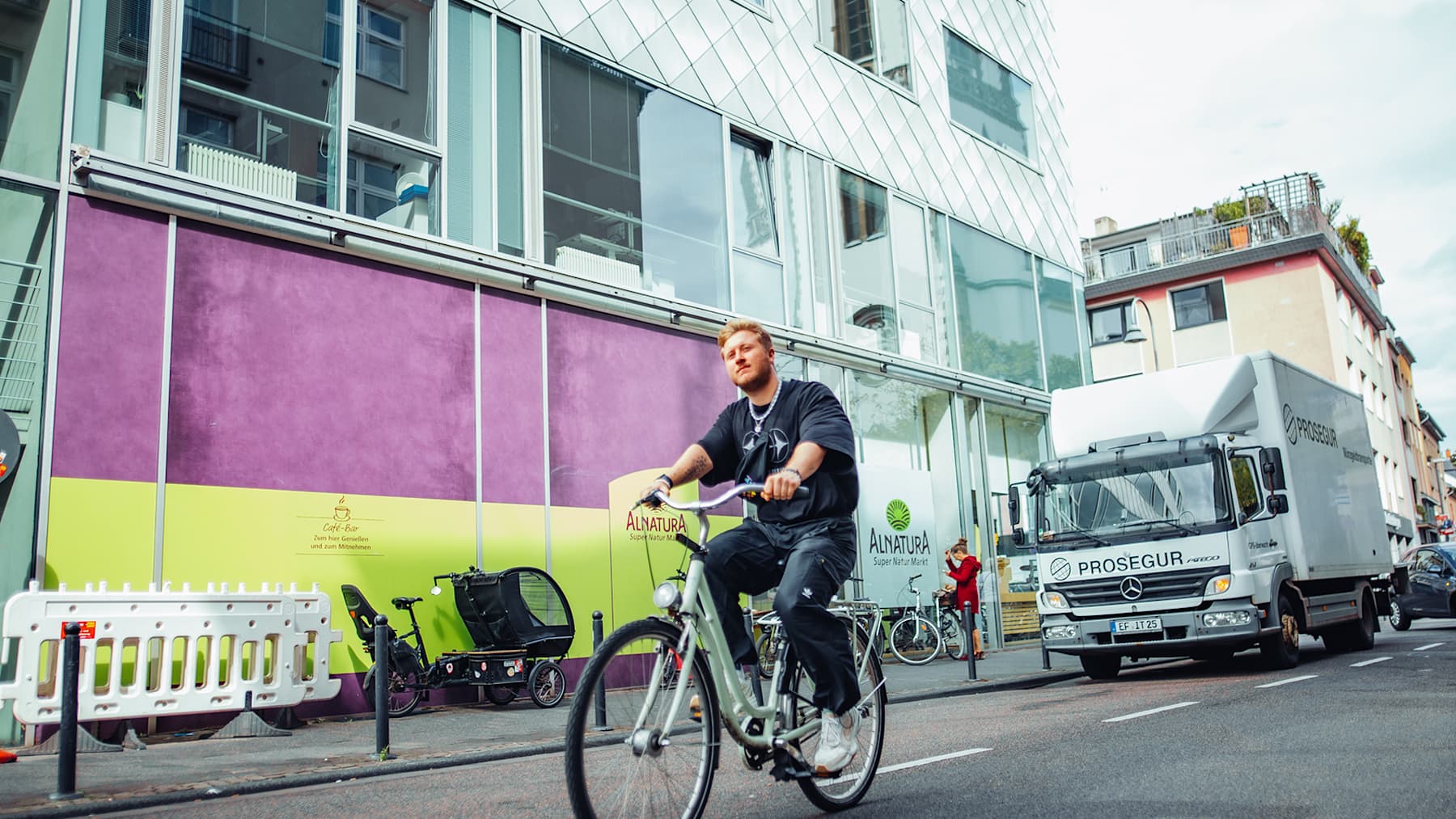 A student is riding his bike down a busy street in Cologne, passing buildings.