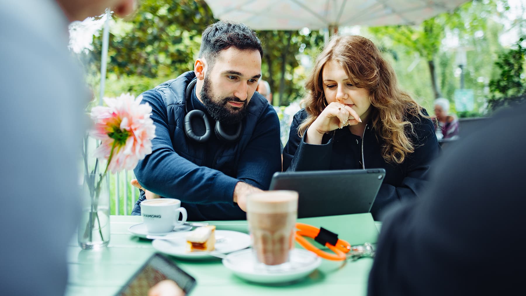 A male student and a female student are intently looking at a tablet while sitting at an outdoor table with coffee and pastries on it.