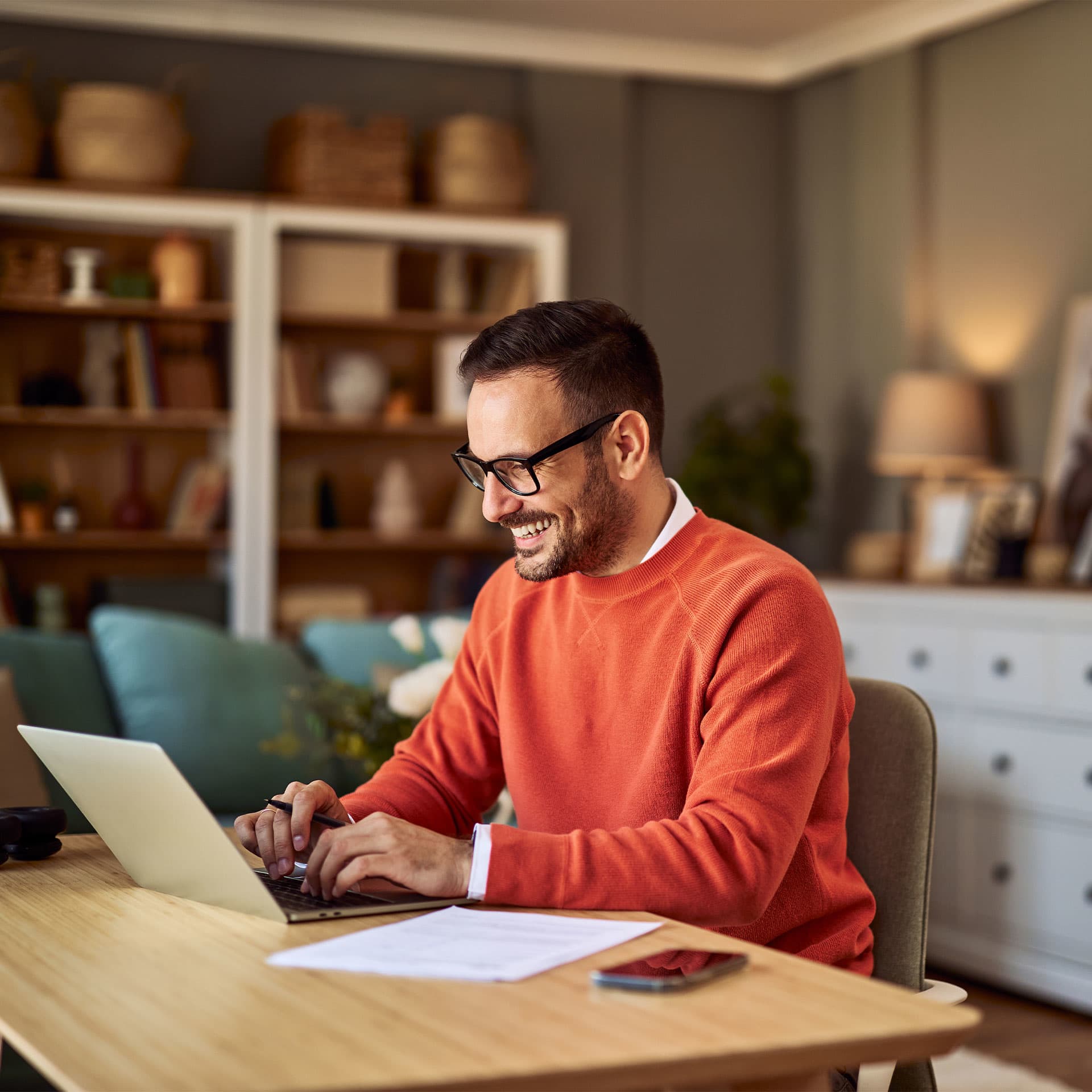 A man wearing an orange sweater and glasses is sitting at his desk, working and smiling.