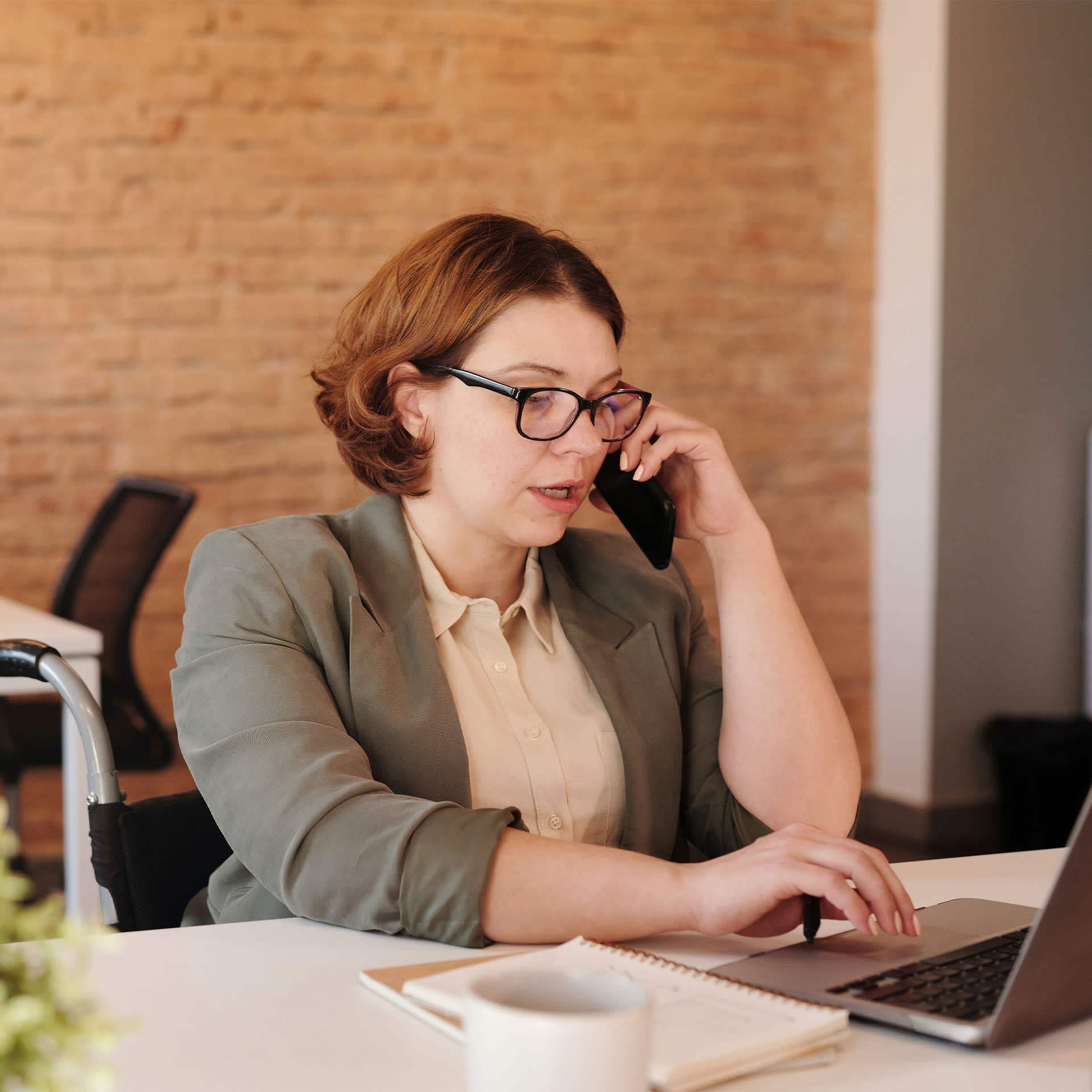 A focused woman multitasks by making a phone call and working on her laptop, focusing on long term care planning in a modern office setting.