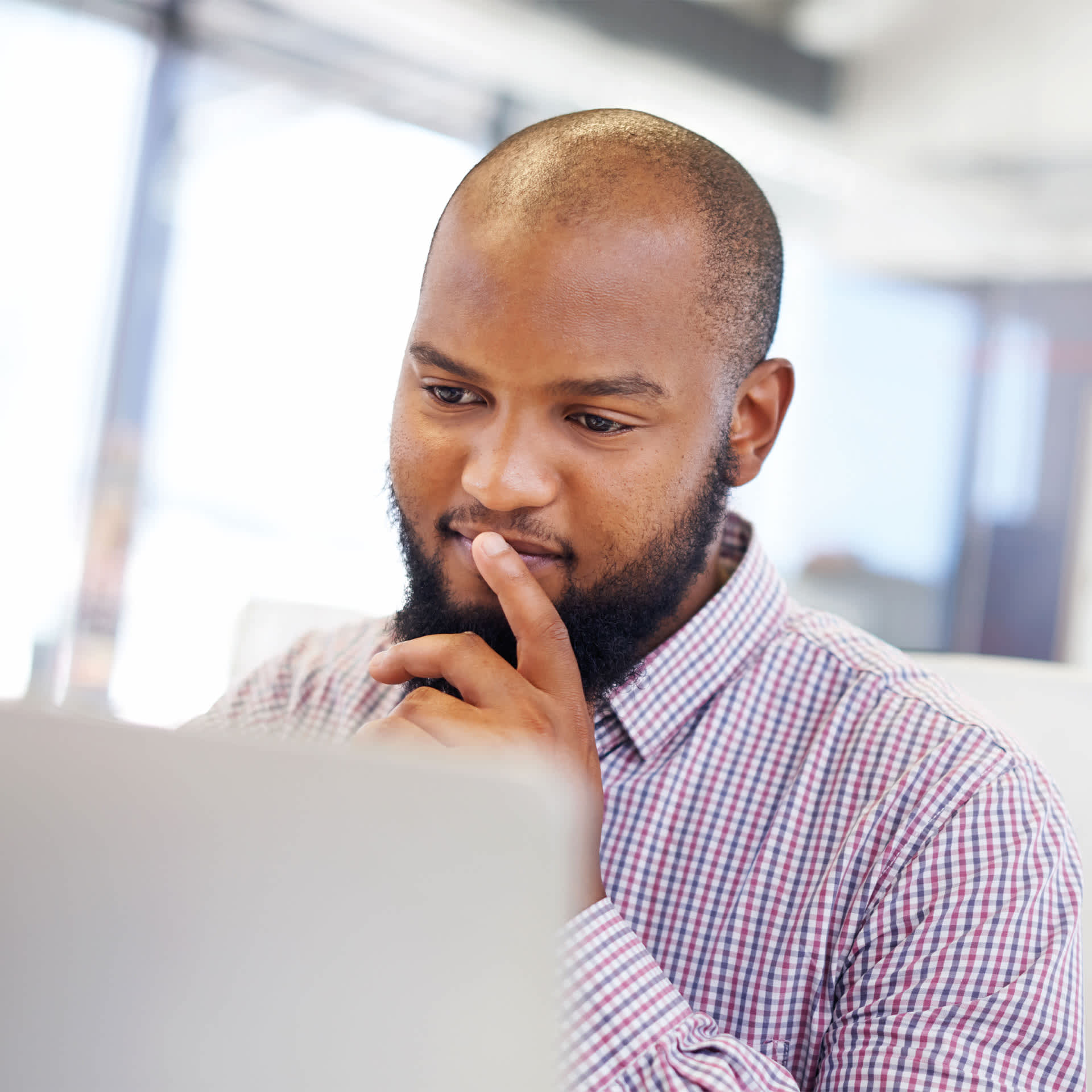 A focused man in a chequered shirt attentively working on his finance diploma on a laptop in an office environment.