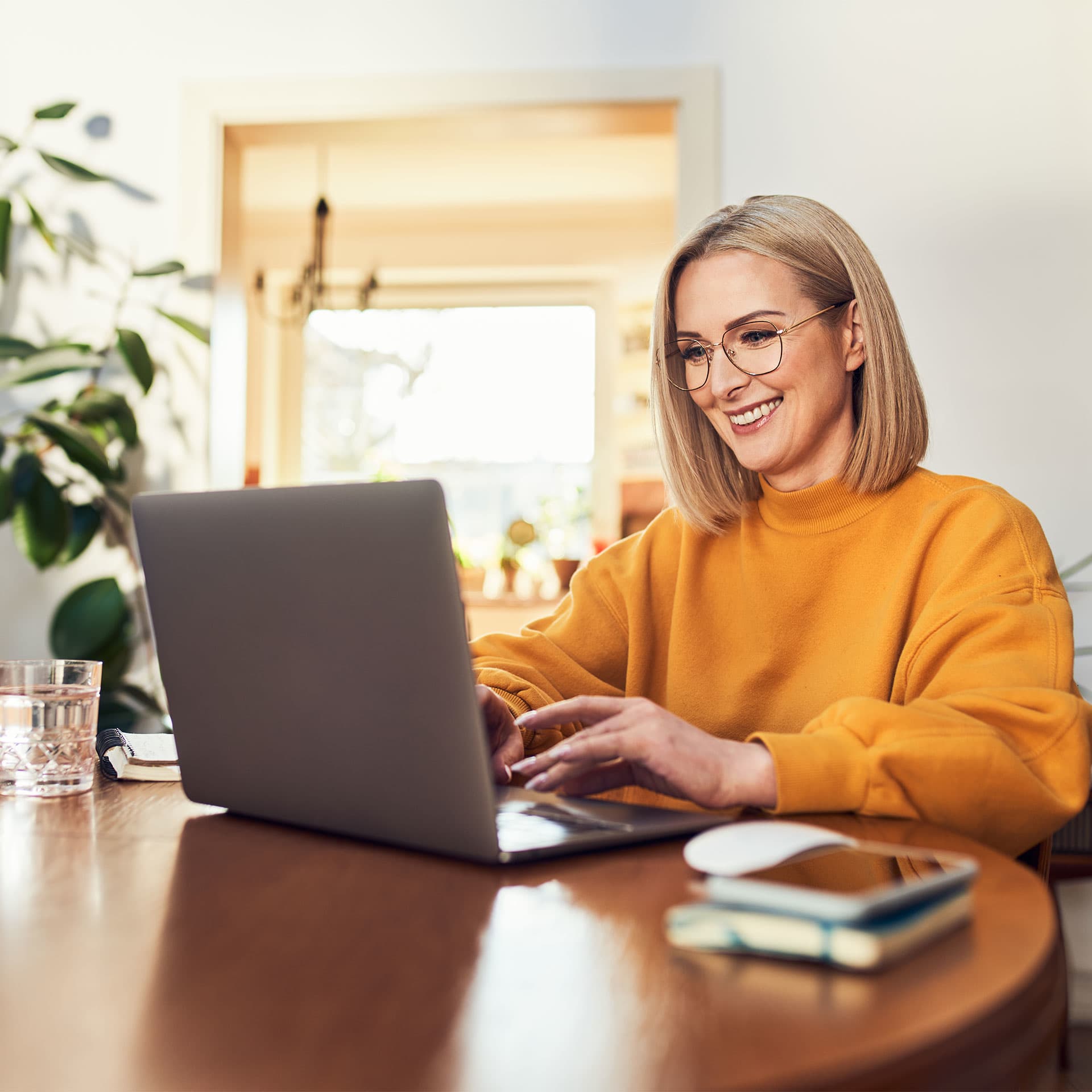 A smiling woman in a yellow sweater working on her paraplanner courses at her laptop in a bright and cosy home office setup.