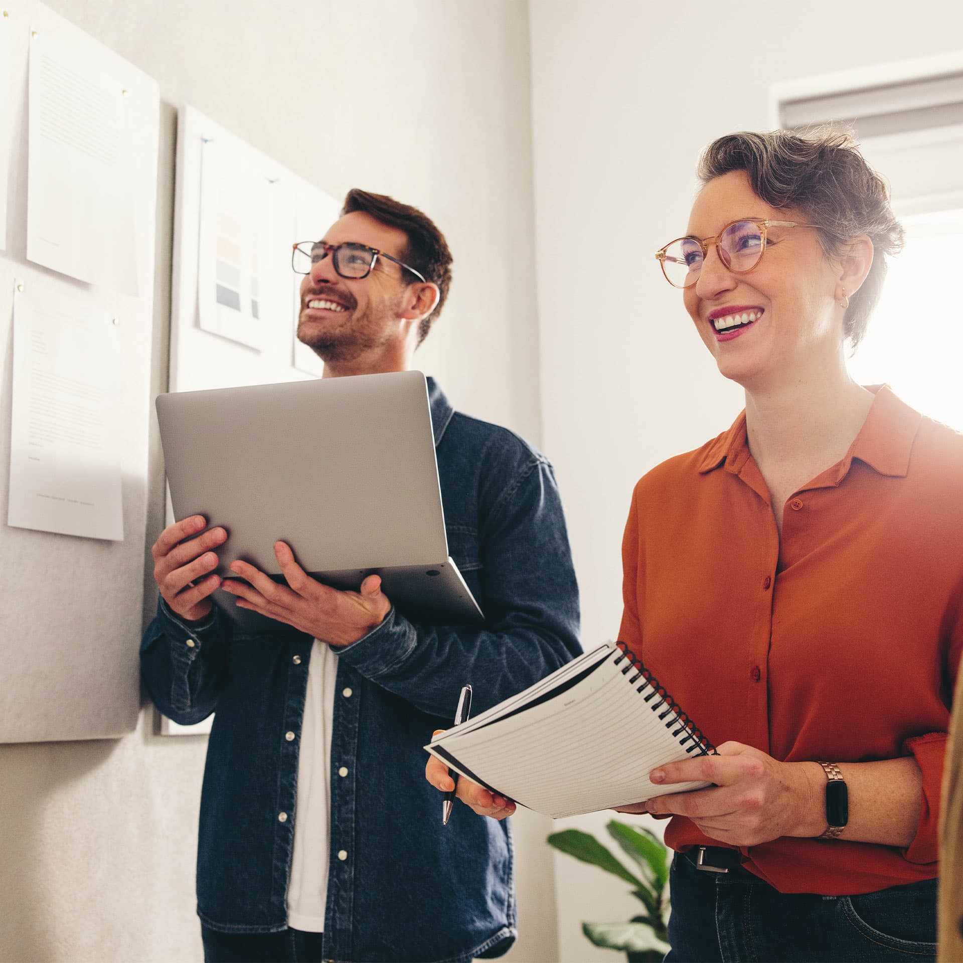 Two professionals sharing a light-hearted moment in a bright office space, one holding a laptop and the other carrying a notebook, discussing about their FSRE module.