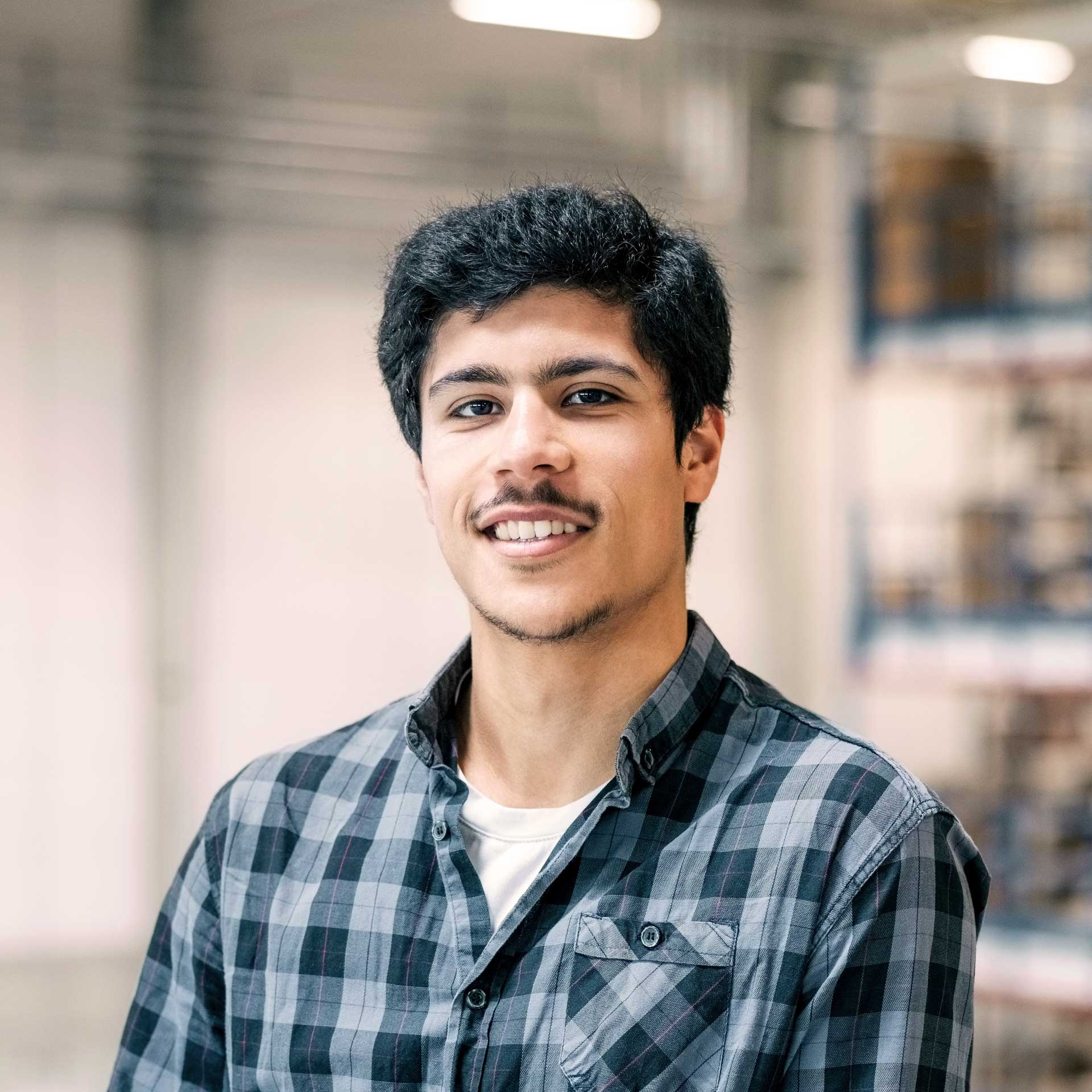 Smiling young student in plaid shirt, standing in a modern, professional indoor setting.