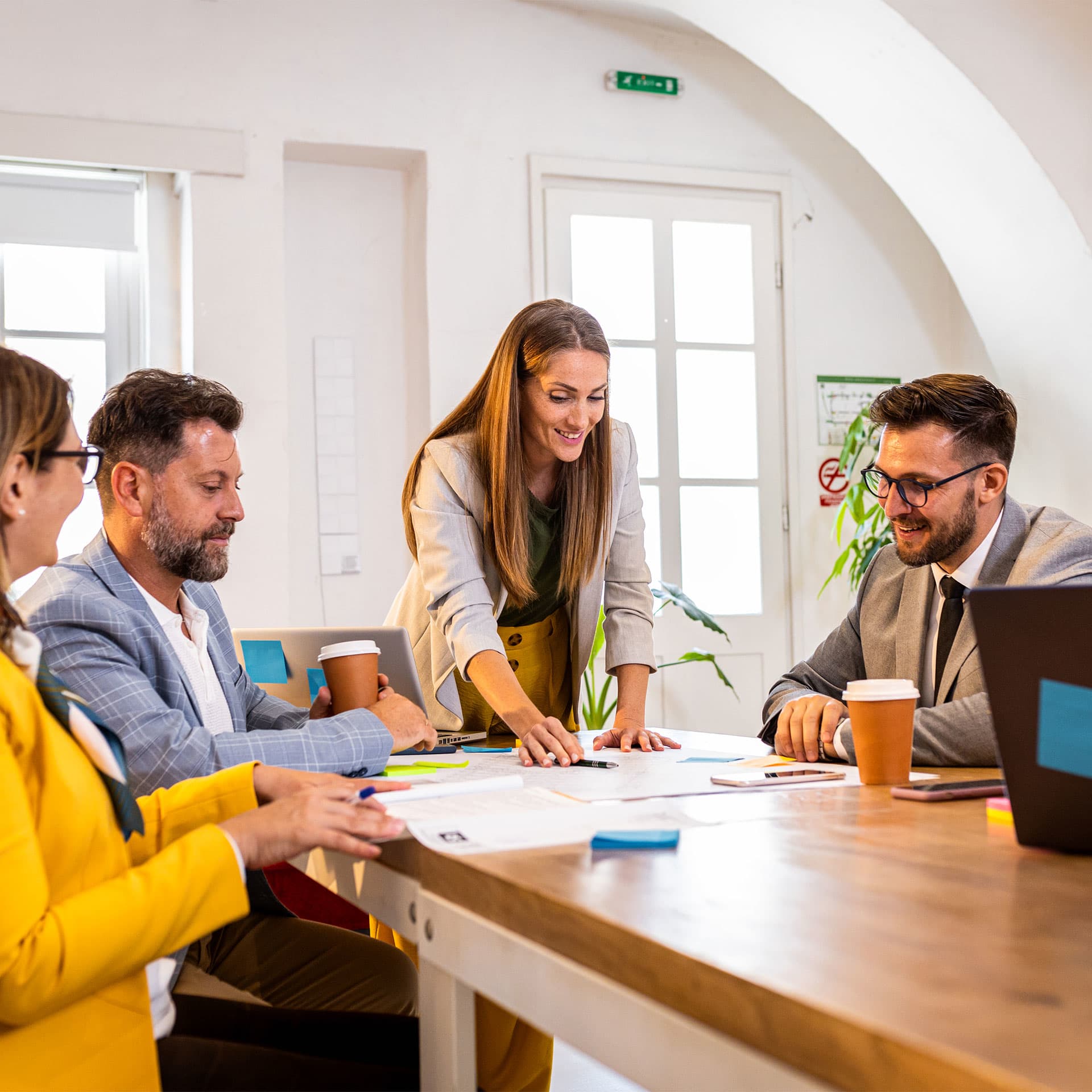 A team of professionals engaged in a collaborative meeting on banking apprenticeships, with one person standing and leading the discussion over documents spread out on a table, while others listen attentively.