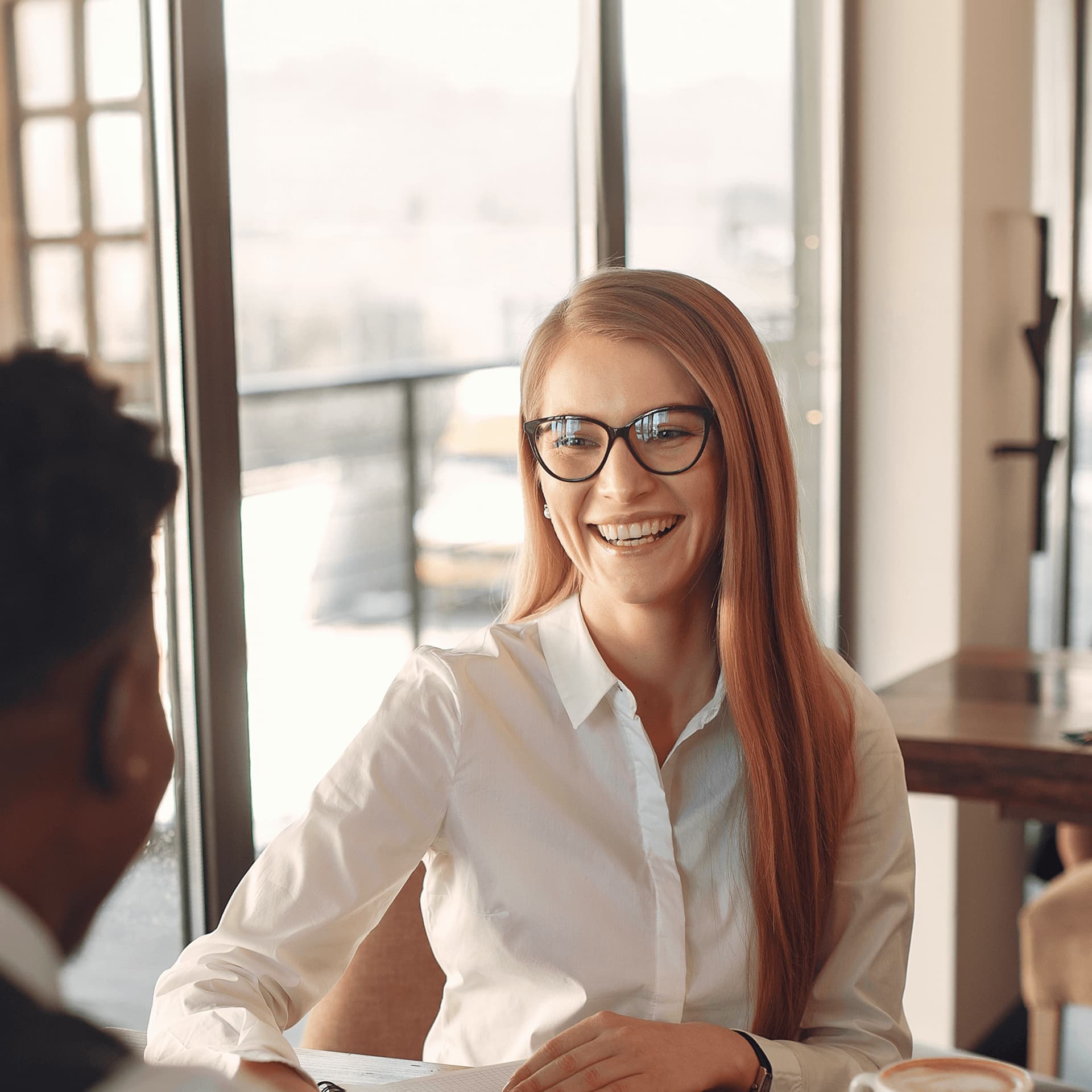 A smiling woman wearing glasses is sat down facing a man, engaging in a conversation about trade finance.