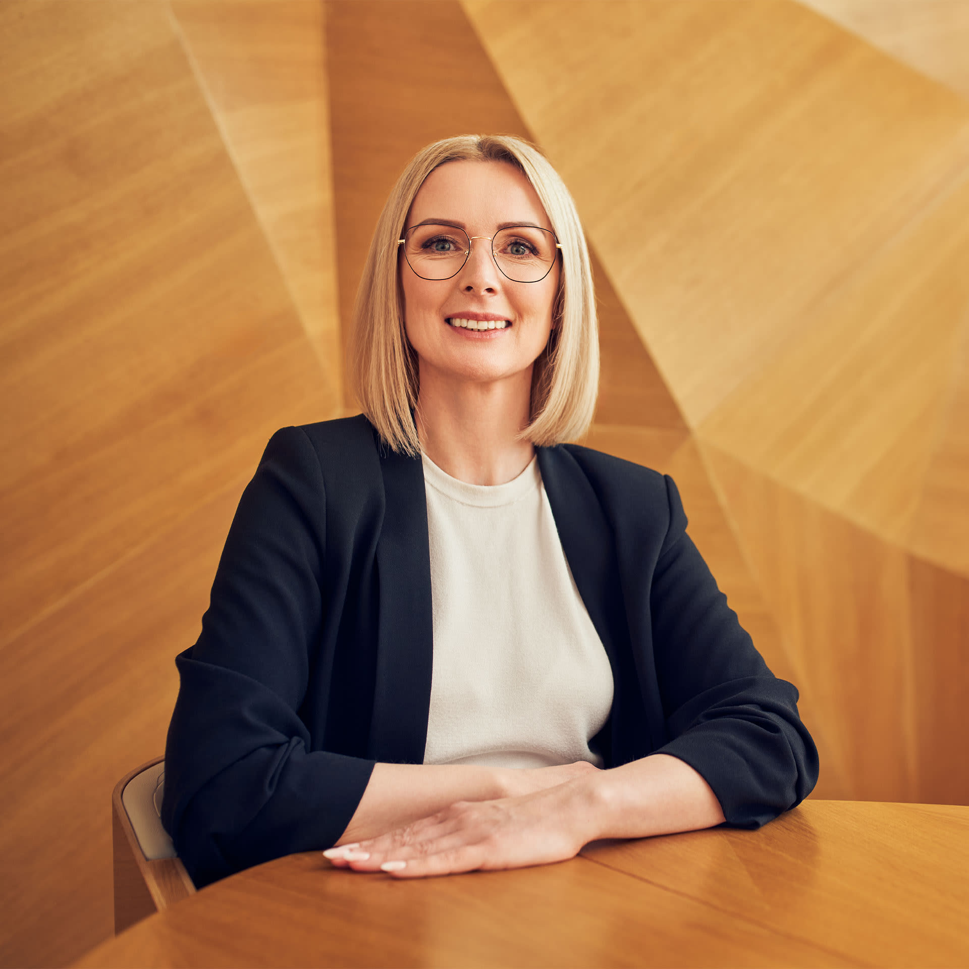 Confident professional woman with a friendly smile sitting at a table against a warm wooden background, specialising in trade finance.