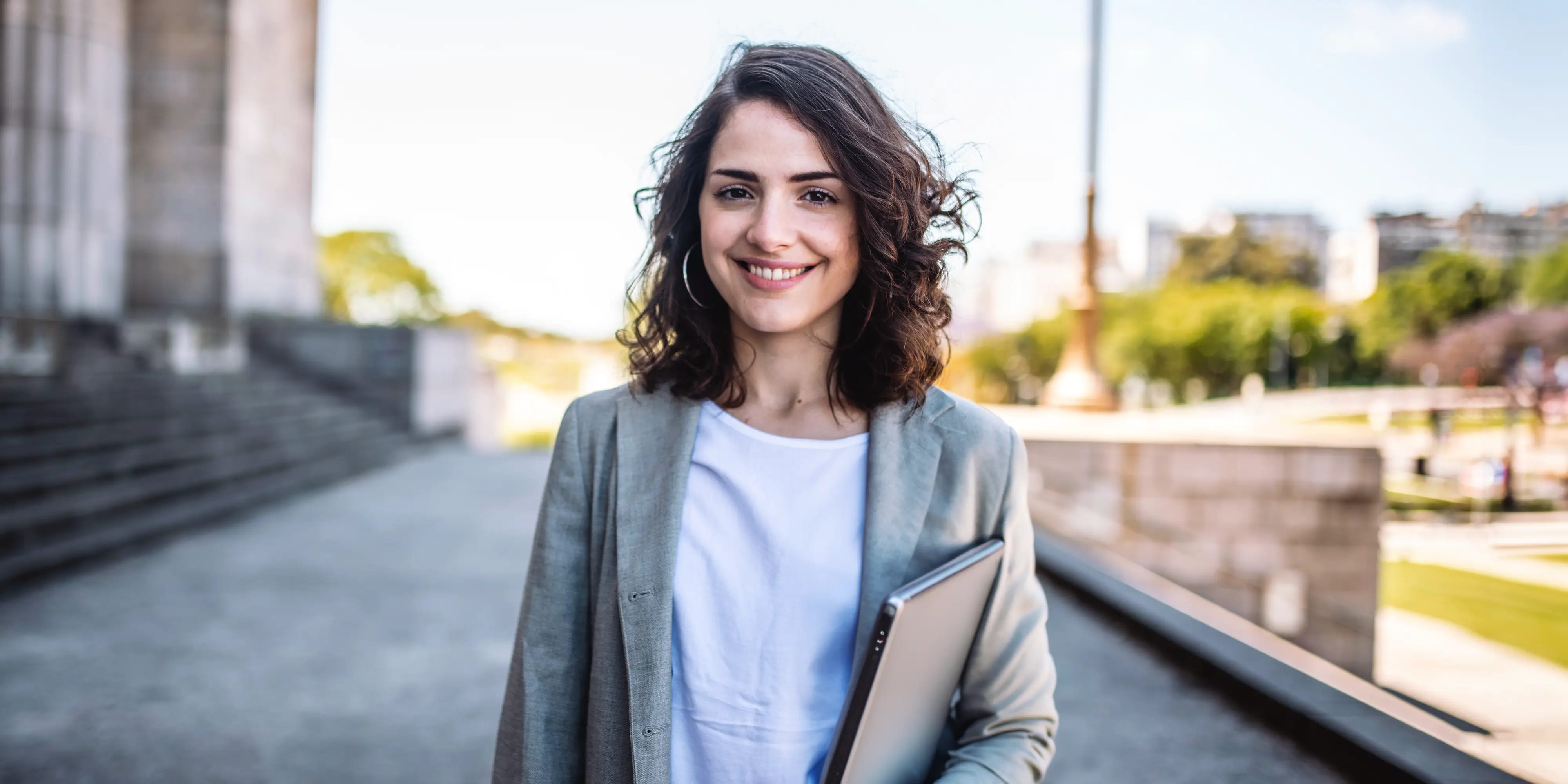 Eine Bachelorstudentin schaut lächelnd in die Kamera und trägt einen Laptop unter ihrem Arm