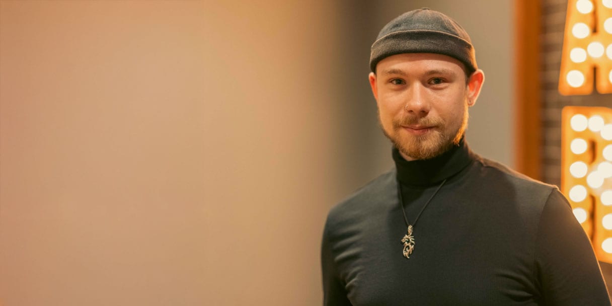 Titelbild zu Artikel "Yannicks Weg: Mediendesign dual studieren und remote arbeiten": Yannick steht lächelnd im Büro von Schenk Immobilien