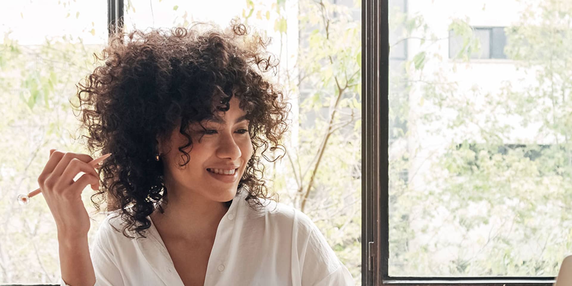 Young woman with curly hair holding a pen and looking at her laptop. On the table there is an open notebook and a mug.