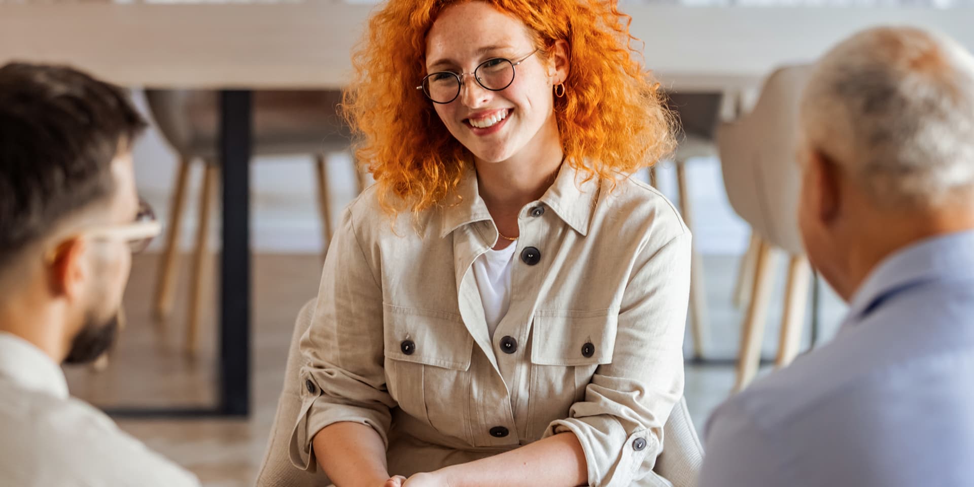 Woman with curly red hair in cream shirt speaks with two men about mental wellbeing in a modern office.