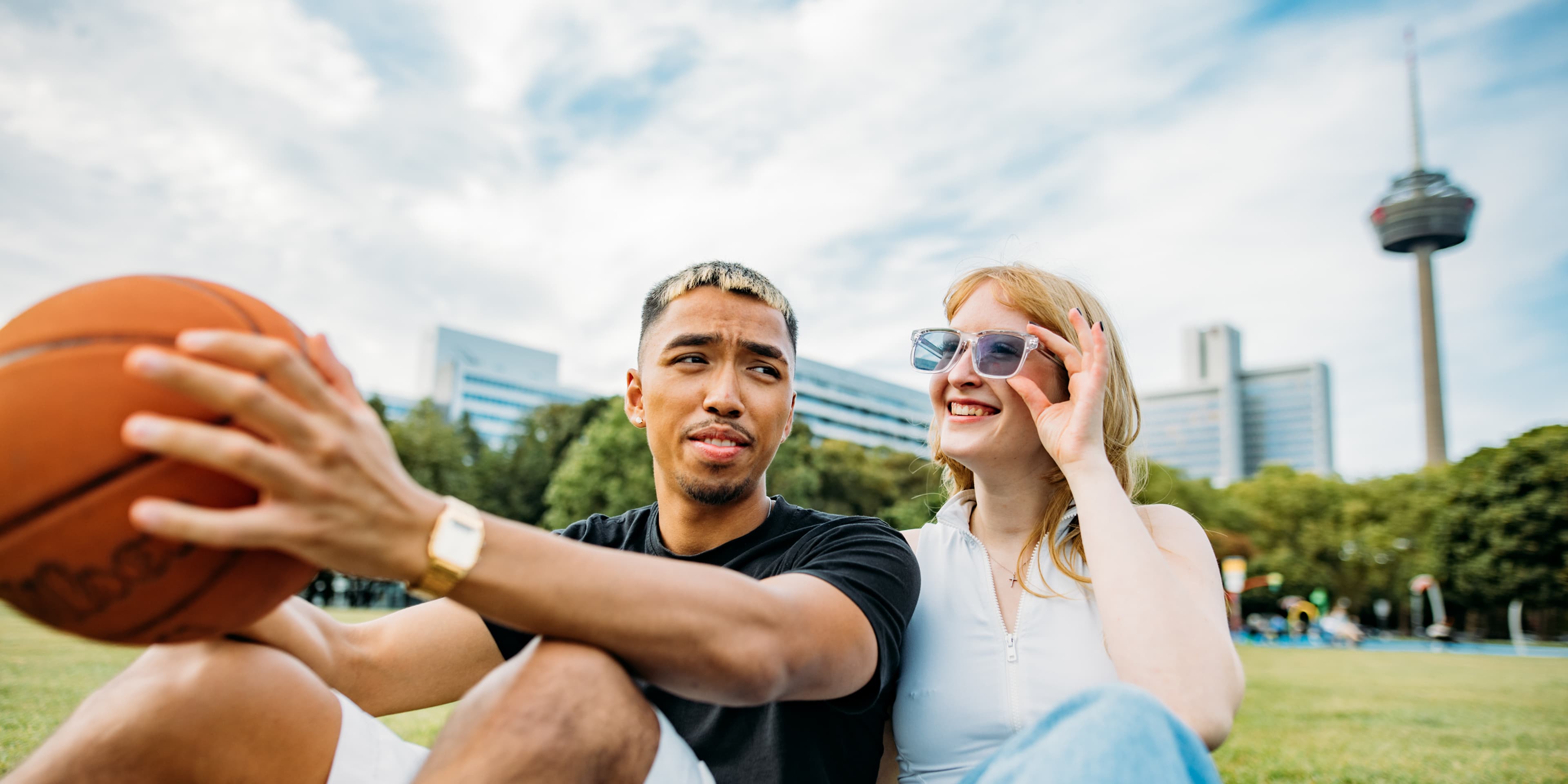 A male student holding a basketball sits on grass next to a female student adjusting her sunglasses, with the Cologne television tower in the background.
