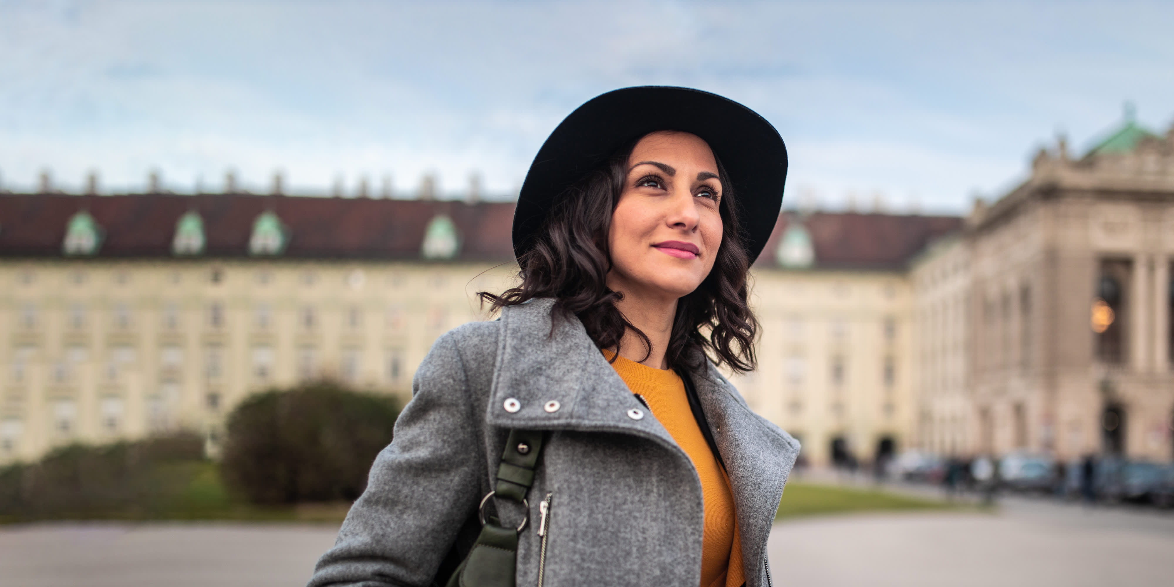 Eine Frau mit Hut und grauem Mantel blickt selbstbewusst in die Ferne. Im Hintergrund ist ein historisches Gebäude zu sehen, das eine städtische Umgebung andeutet. Die Atmosphäre wirkt ruhig und stilvoll.