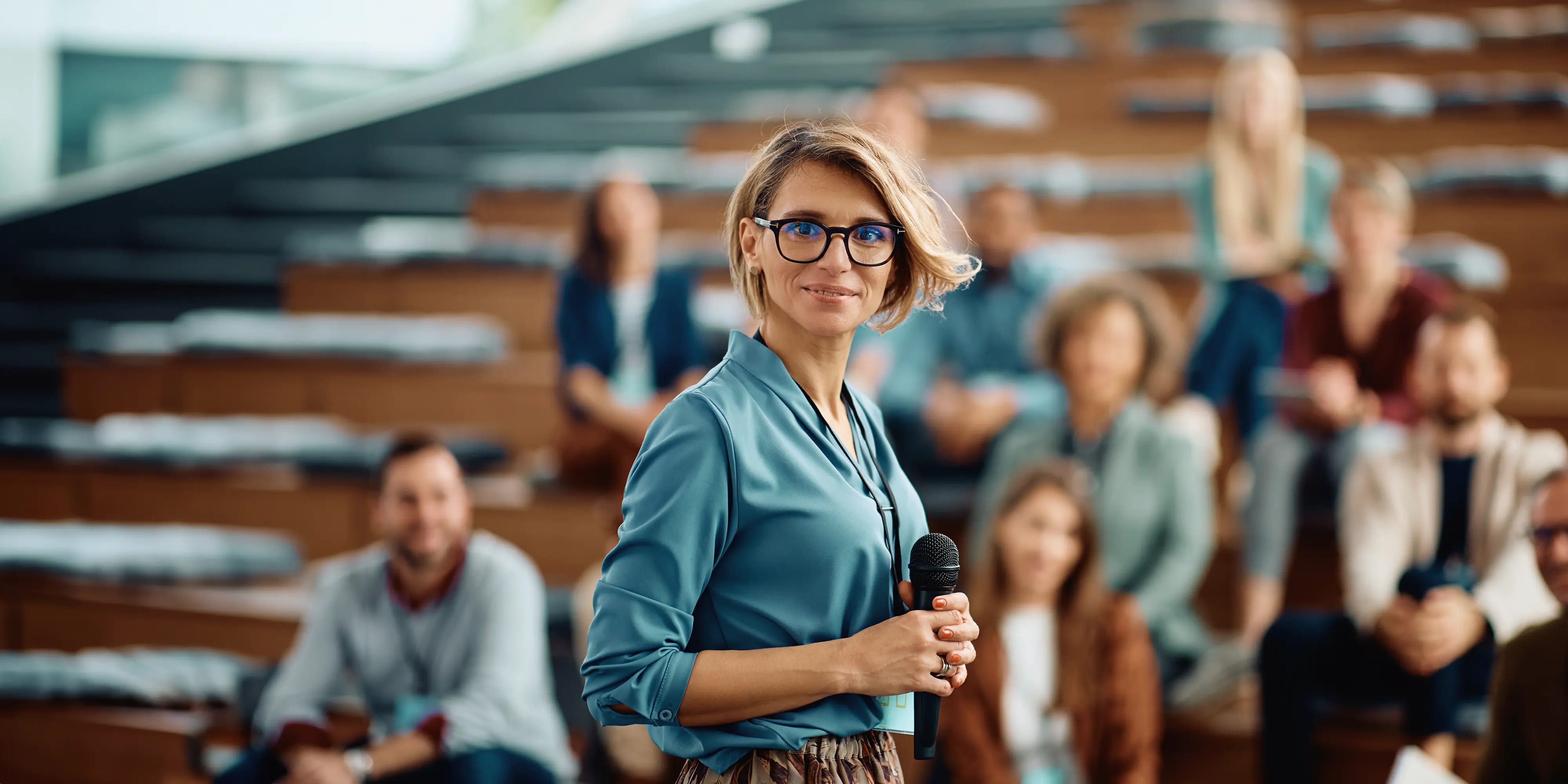 Fachtagung ISPC: Eine Frau hält vor einem Publikum ein Mikrofon in der Hand und schaut in die Kamera