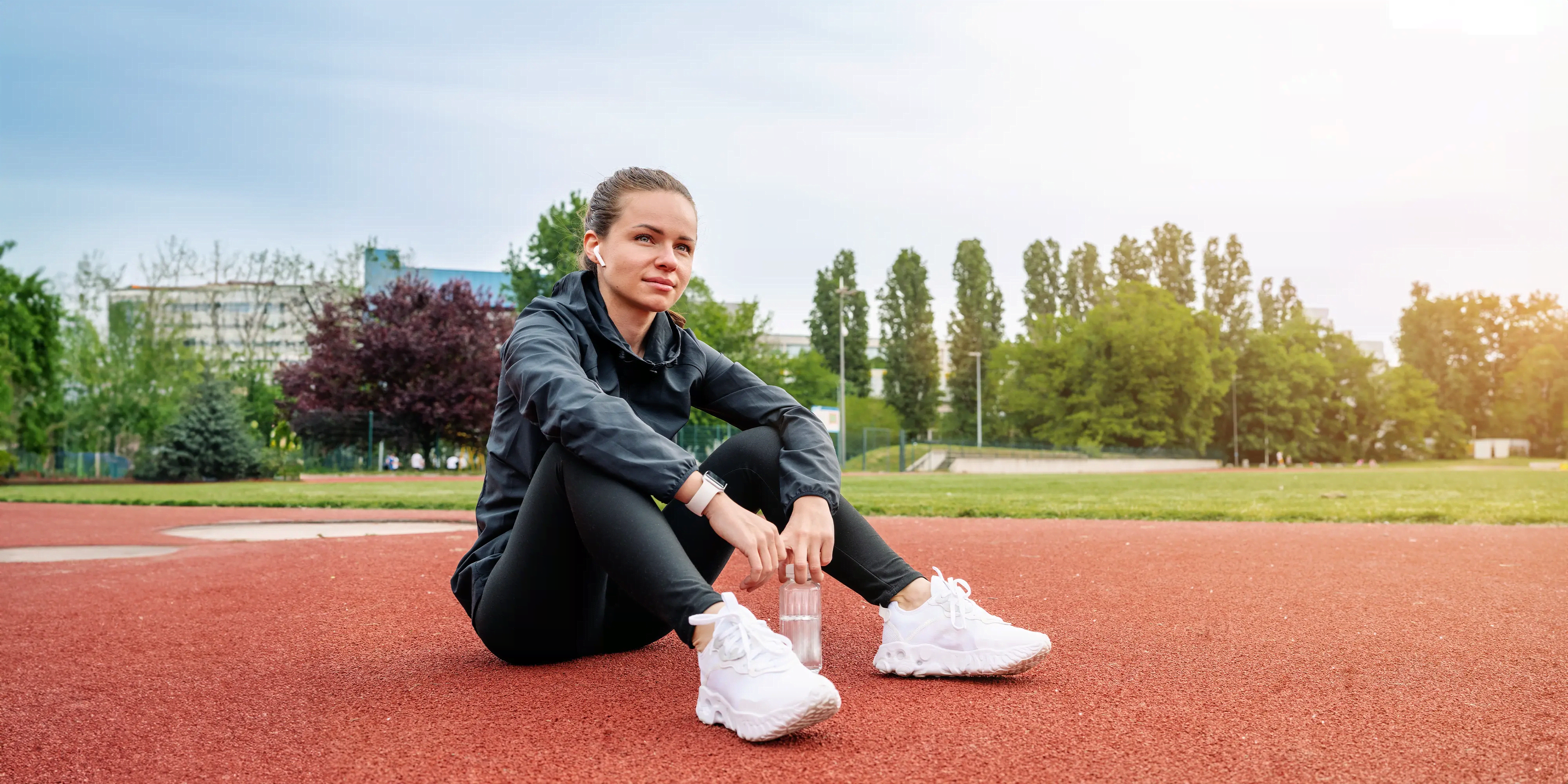 Titelbild der Infoseite "Sportpsychologie berufsbegleitend": Studentin sitzt auf dem Boden eines Sportplatzes mit Wasserflasche und hört Musik