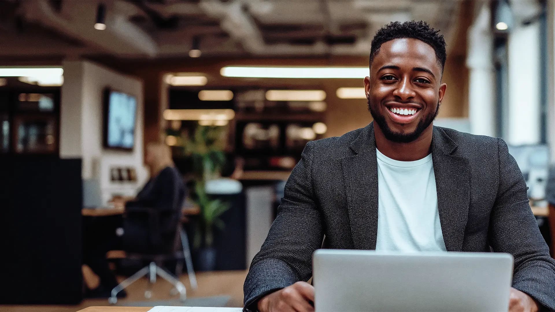 Titelbild zum Artikel: "Was macht ein:e E-Commerce Manager:in?" Ein E-Commerce Manager sitzt am Laptop und schaut lächelnd in die Kamera, im Hintergund sieht man ein Büro