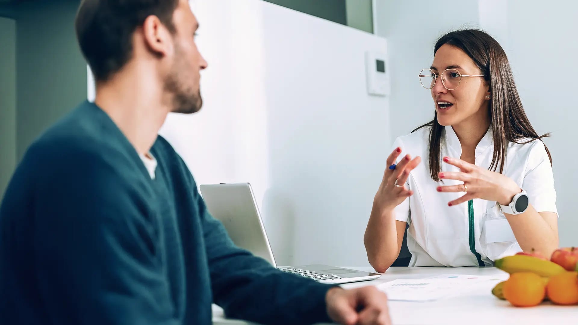Titelbild zum Artikel: "Ernährungsberater werden": Eine Frau im weißen Kittel spricht gestikulierend mit einem Klienten am Tisch