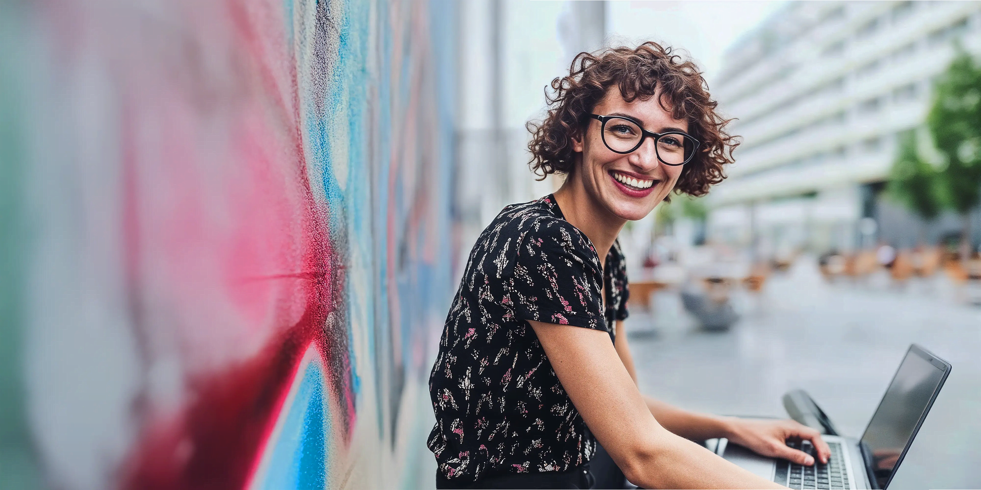Studentin arbeitet lächelnd mit Laptop im Freien vor einer bunten Wand