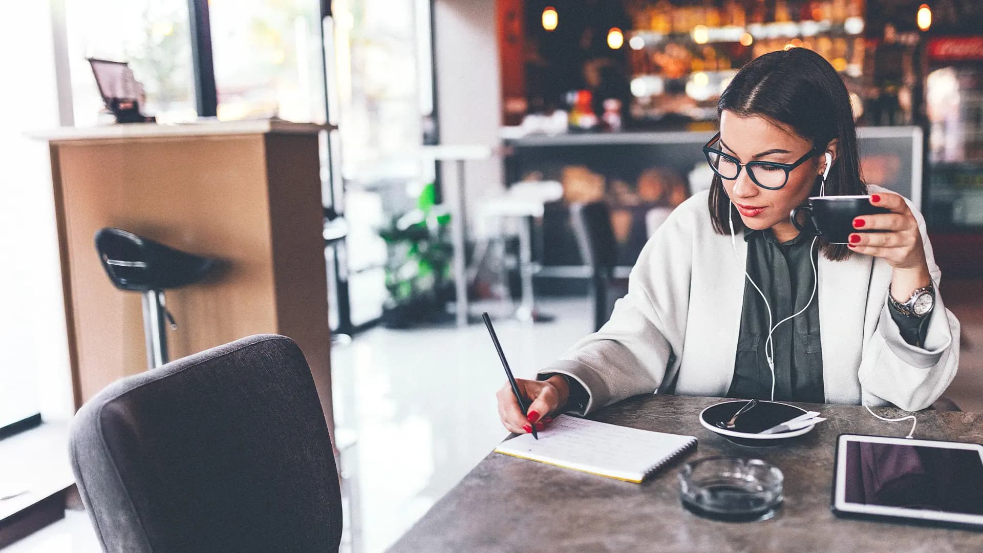 Titelbild zum Artikel "Draußen Lernen: 5 Lieblingsorte": Studentin lernt draußen im Café mit Notizen, Tablet und Kaffee.