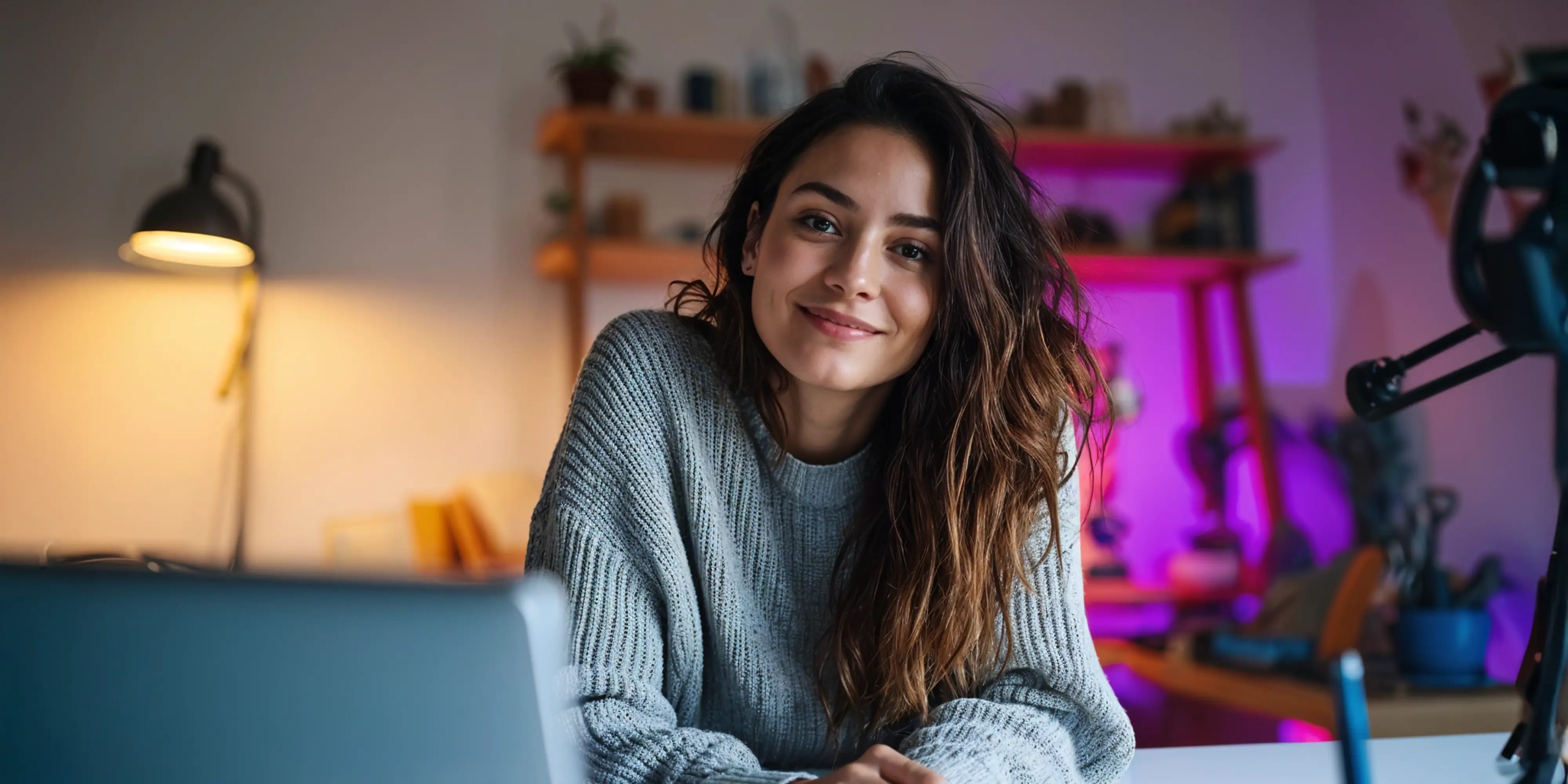 Studentin lächelt in einer gemütlichen Wohnung und moderner Beleuchtung, vor ihr steht ein Laptop